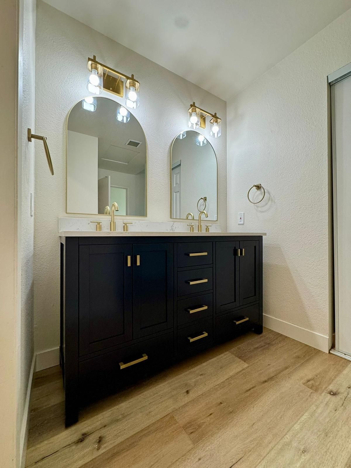 Black double vanity with gold fixtures, two arched mirrors, light-colored walls and wood-look flooring.