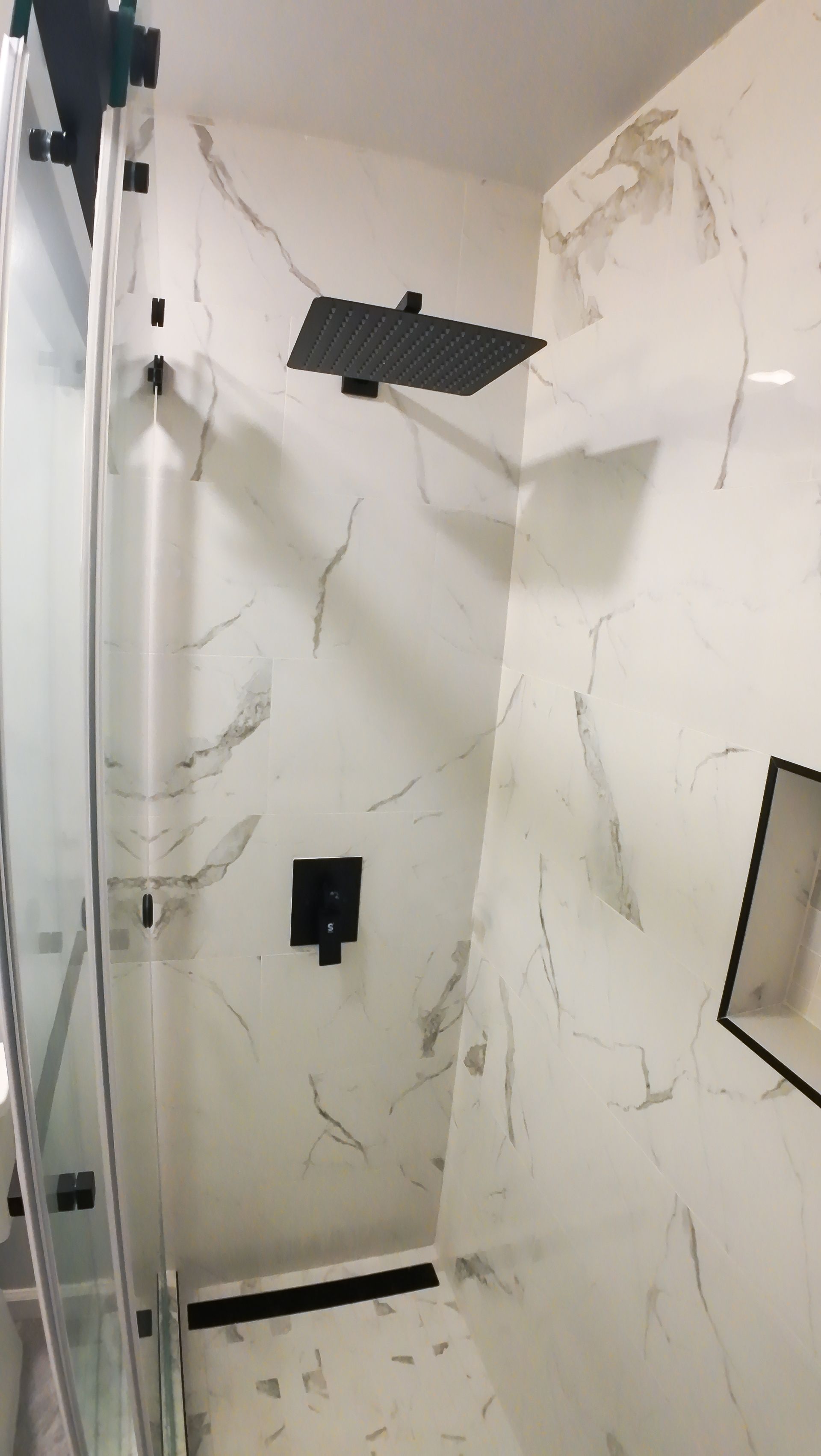 Modern shower with black fixtures and white marble-look tile. Glass door and recessed shelf.
