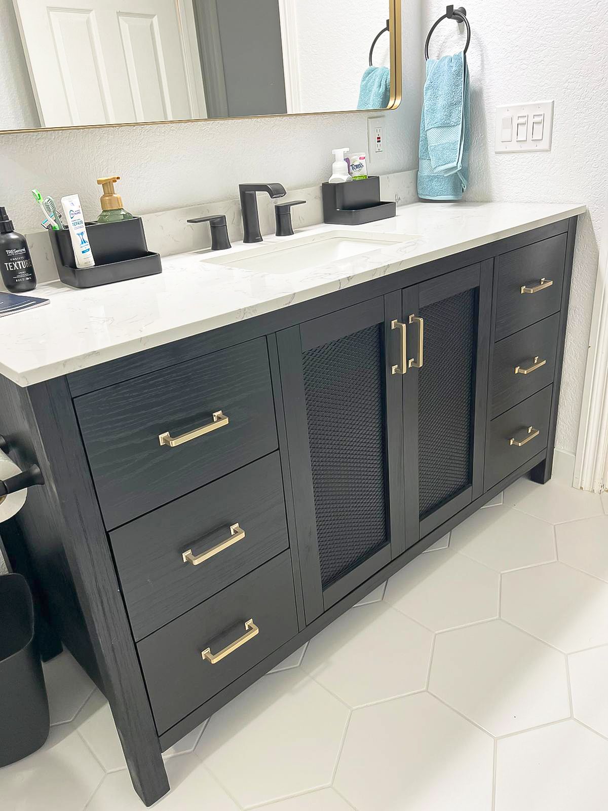 Black bathroom vanity with gold hardware and mesh front doors; white countertop and hexagonal floor tiles.