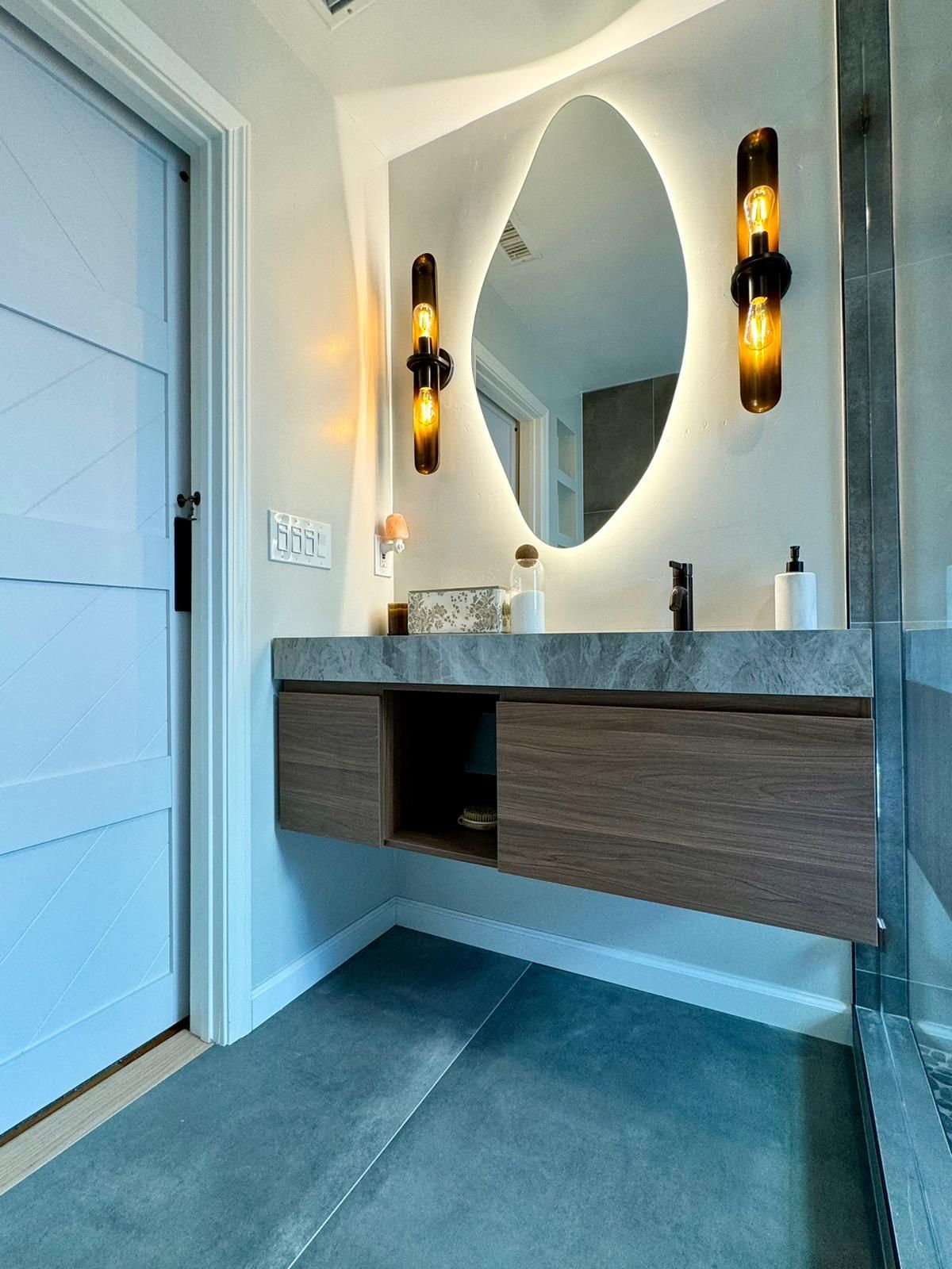 Modern bathroom with floating wooden vanity, oval backlit mirror, sconces, and light blue door.