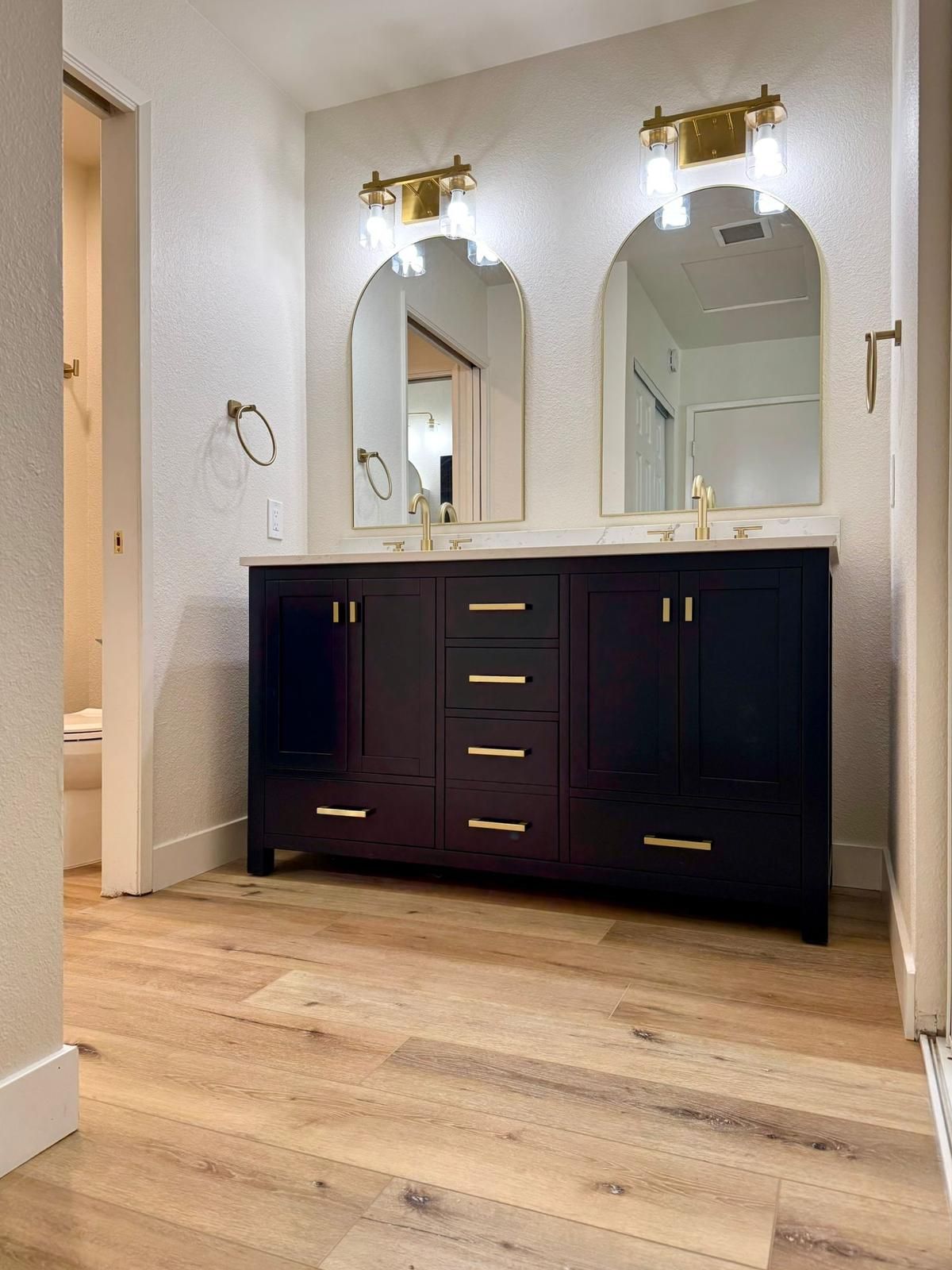 Bathroom with navy vanity, gold hardware, arched mirrors, and wood-look flooring.