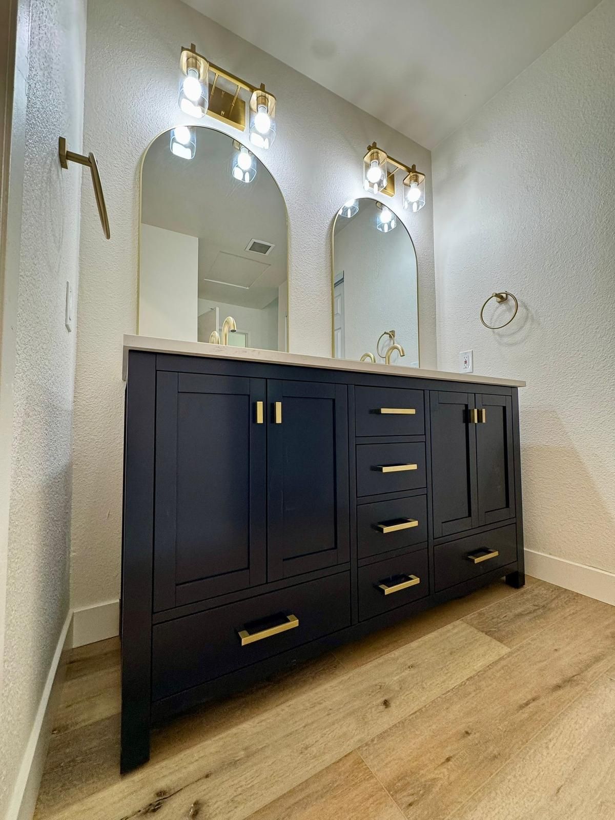 Navy blue bathroom vanity with gold hardware, arched mirrors, and warm wood flooring.