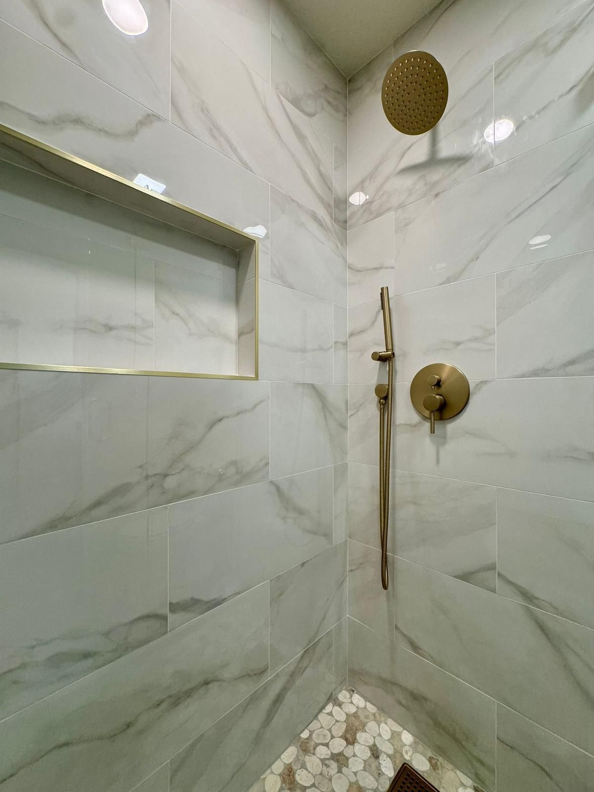 Shower with white marble-look tiles, gold fixtures, built-in shelf, and pebble floor.