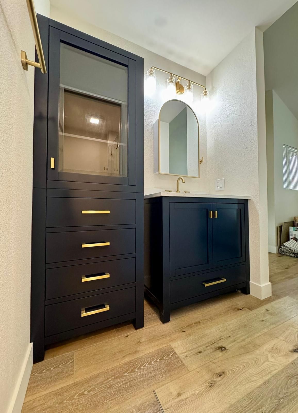 Navy blue bathroom cabinetry with gold hardware, arched mirror, and wood flooring.