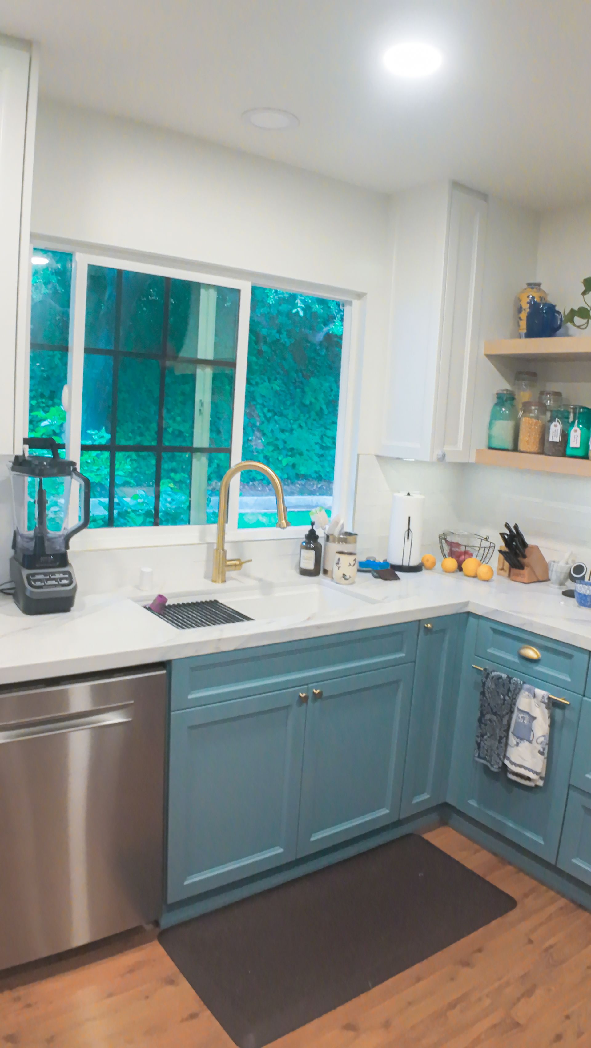 Blue kitchen cabinets, gold faucet, stainless steel appliances, and a window overlooking greenery.