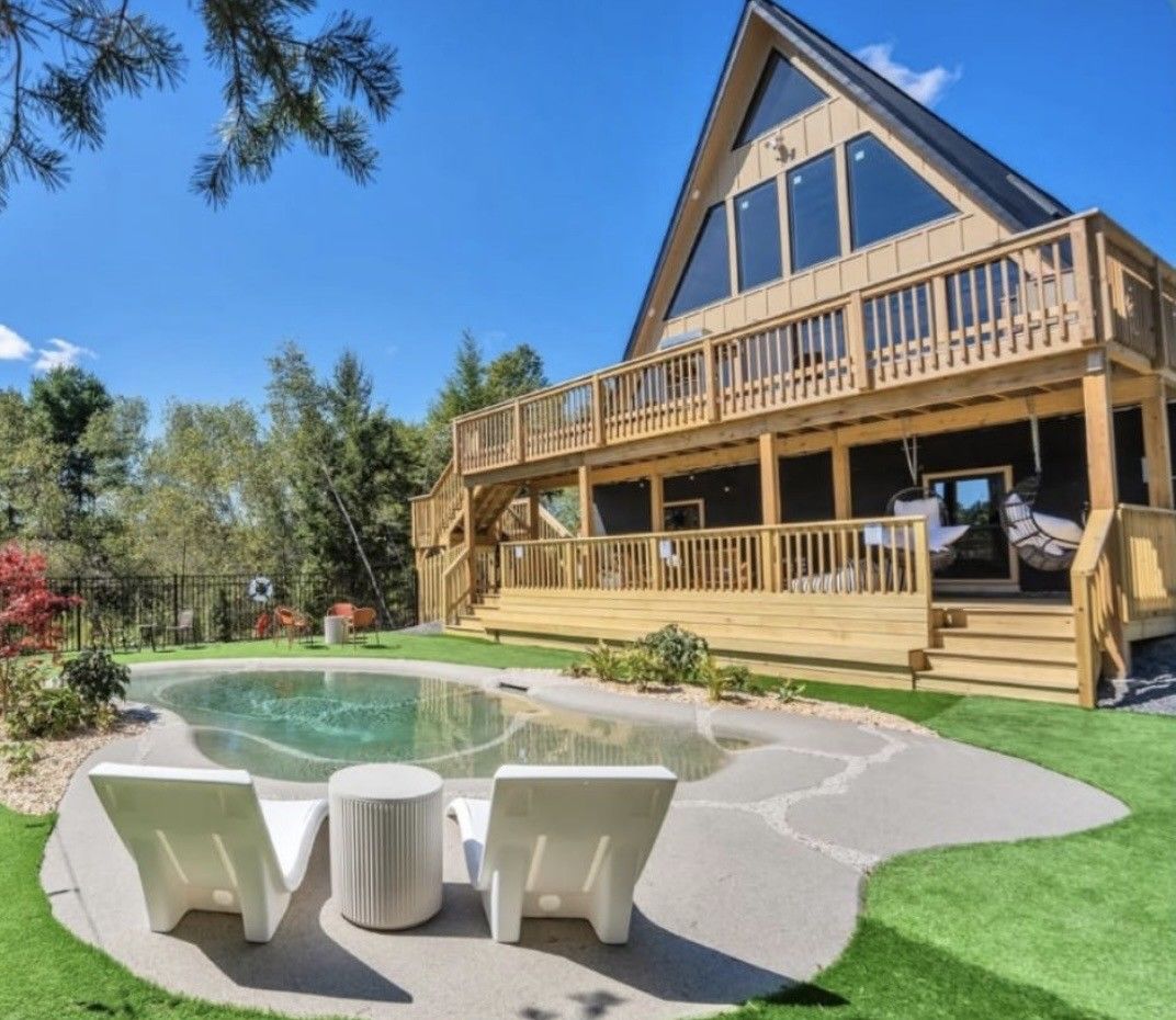 A-frame house with a wooden deck overlooking a pool. Two white lounge chairs face the water on a sunny day.
