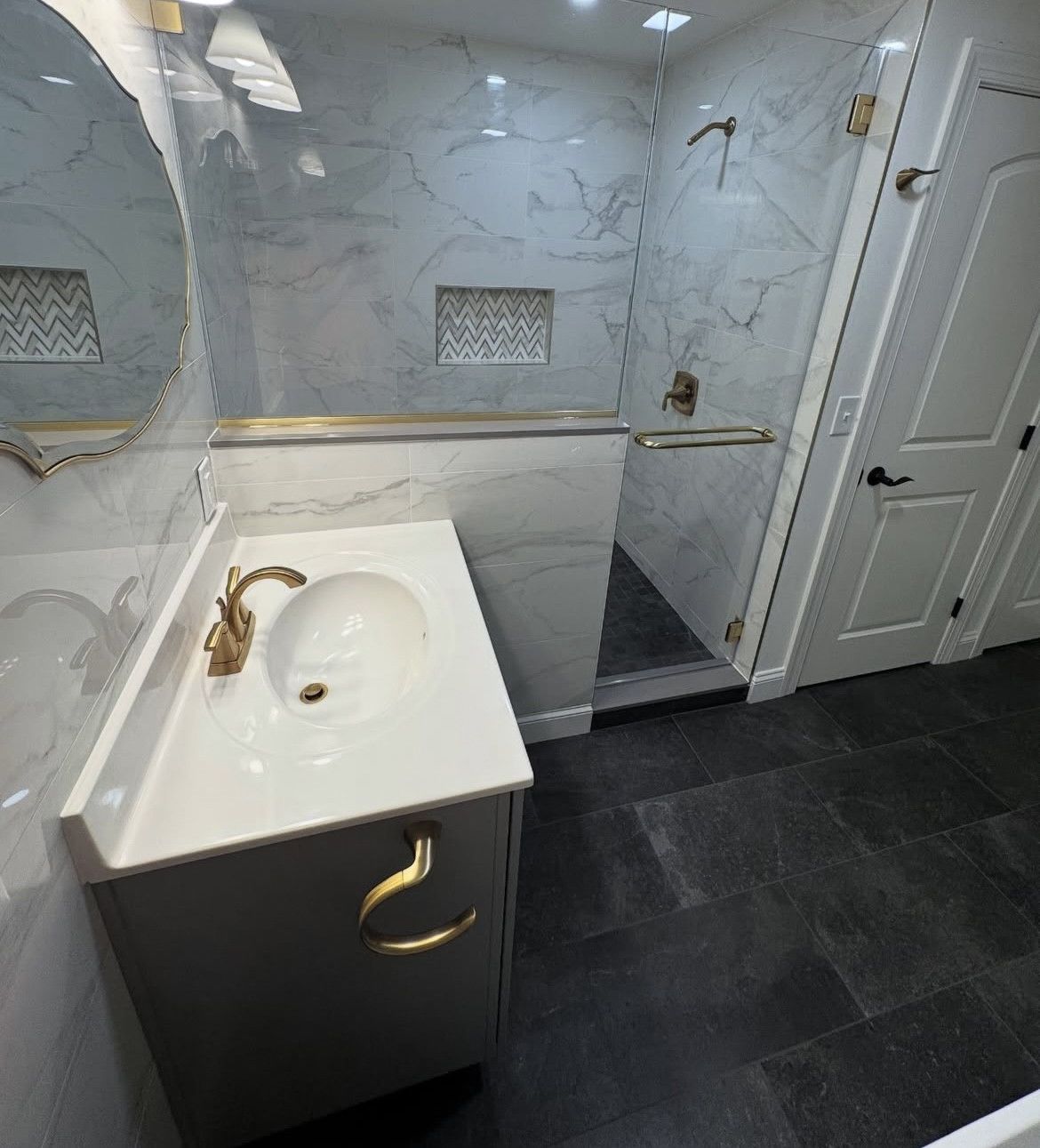 Modern bathroom with white and gray marble tile, glass shower, gold fixtures, and dark floor.