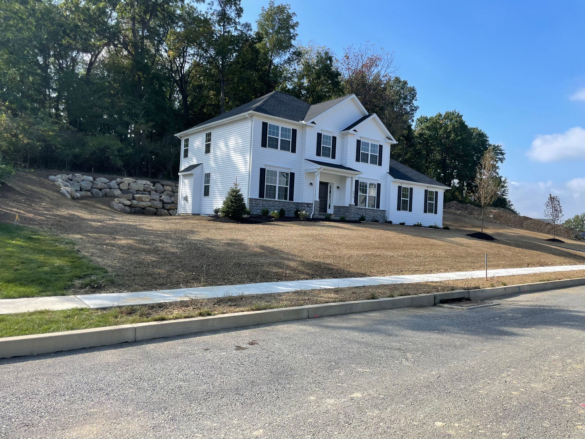 White two-story house with black shutters, on a sloped grassy lot, with a retaining wall and trees in the background.