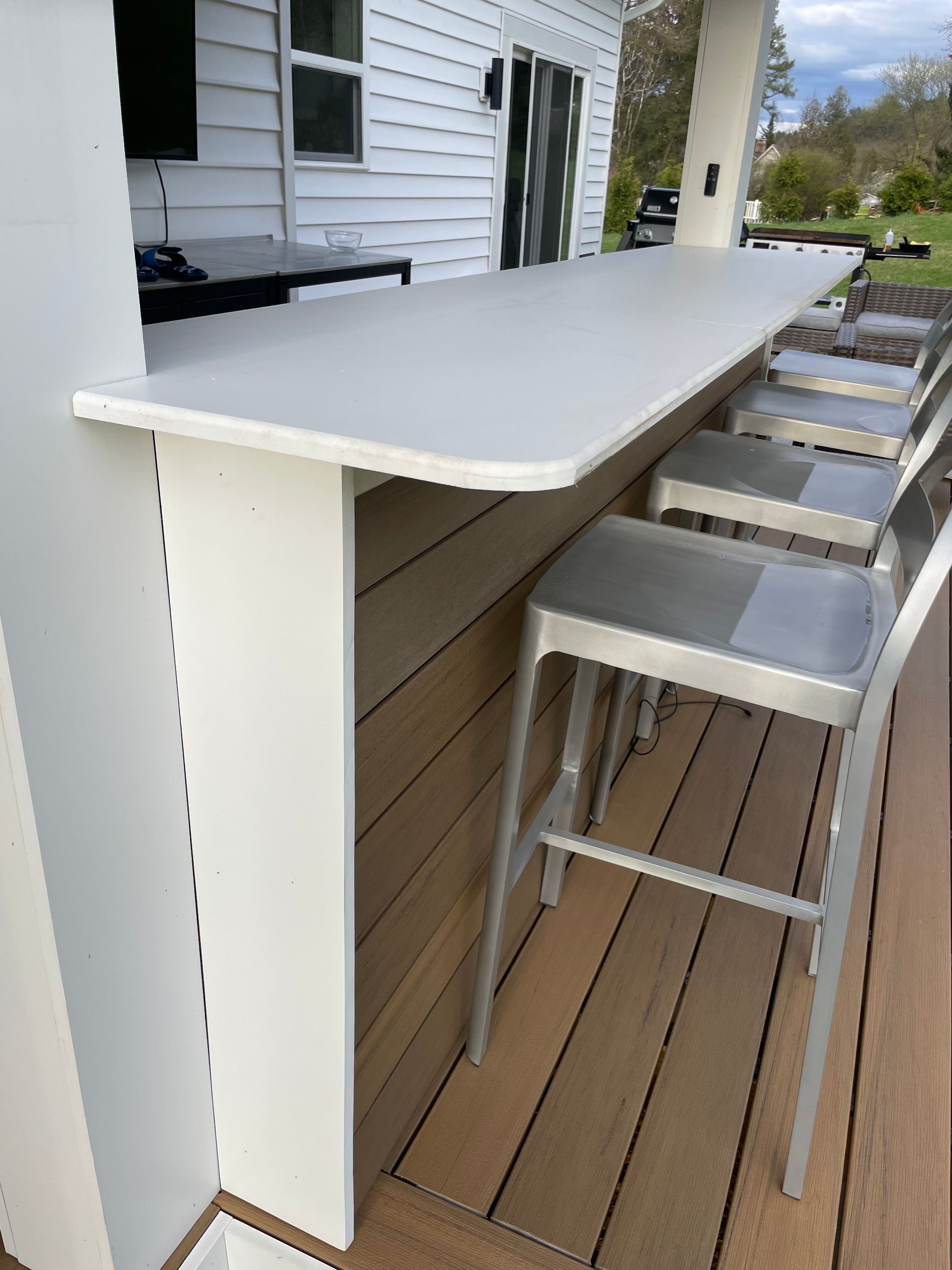 Outdoor bar with white countertop, metal stools, and wood paneling on a deck.