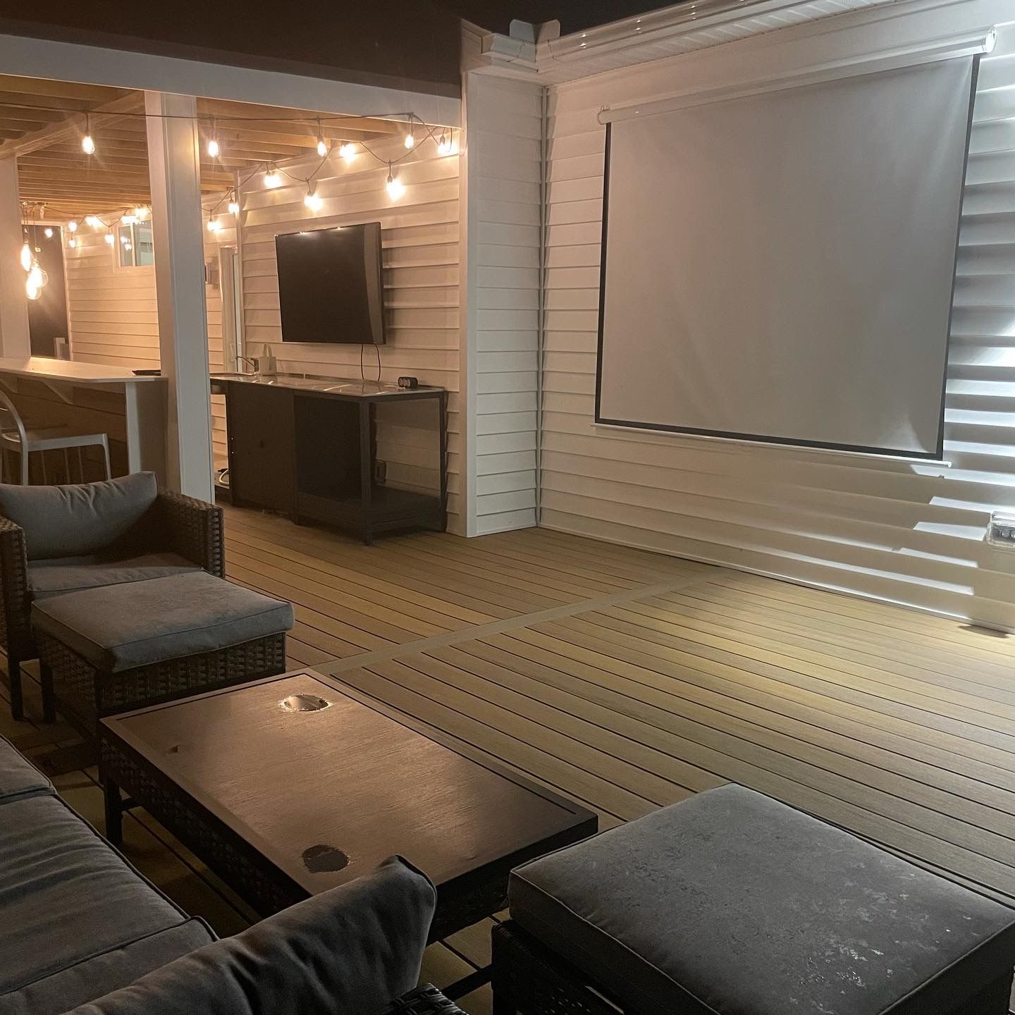 Outdoor entertainment area with a projector screen, TV, and seating, lit by string lights.