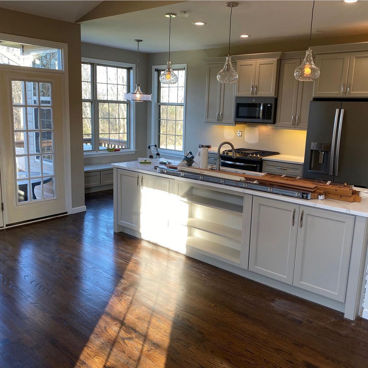 Kitchen with light gray cabinets, island, and dark wood floors. Windows let in sunlight.