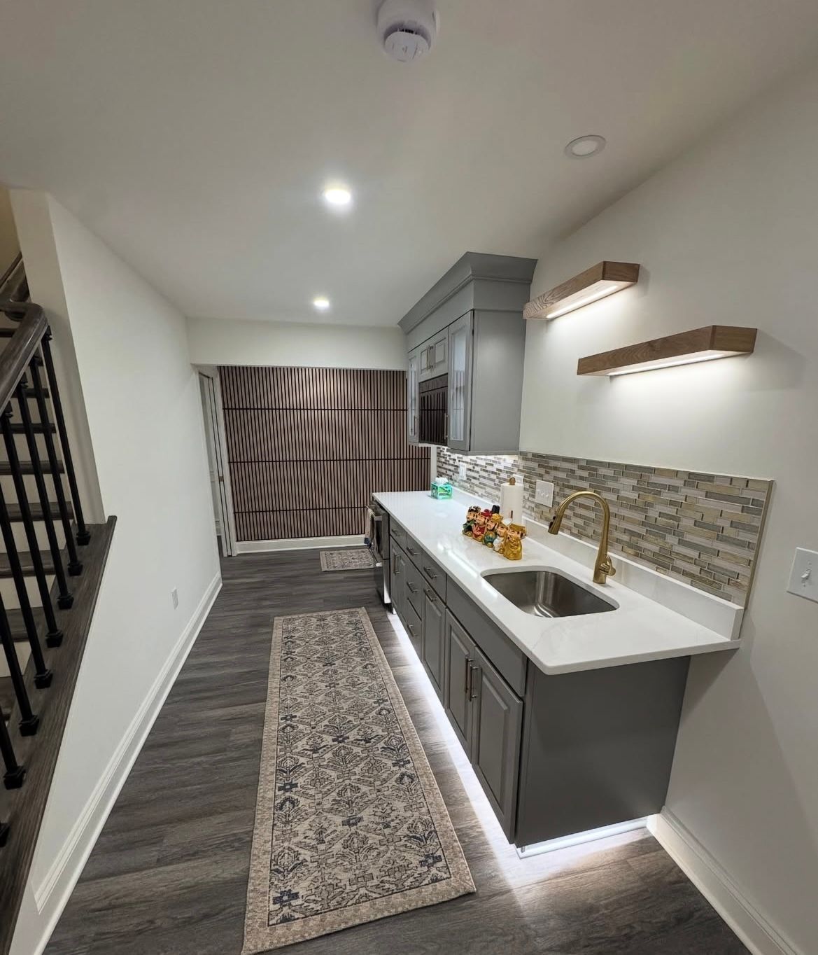 Basement bar with gray cabinets, white countertop, sink, and decorative backsplash. Rug and under-cabinet lighting.