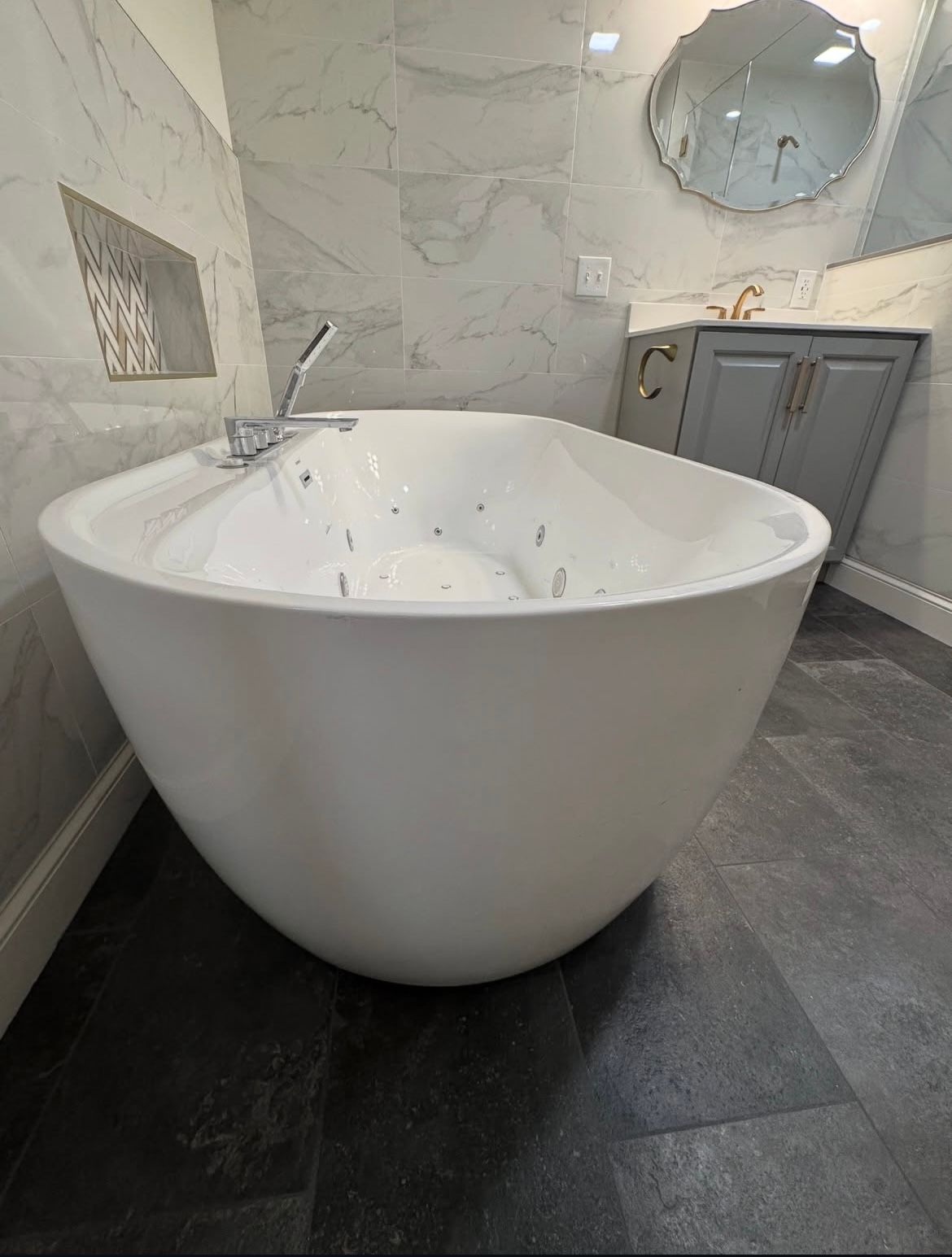 White, egg-shaped bathtub in a modern bathroom. Gray tile floor and marble-look wall tiles.