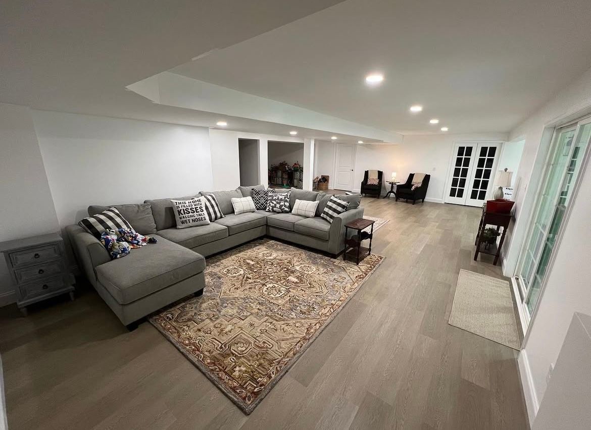 Spacious basement with sectional sofa, rug, and light wood flooring. Several chairs are in the background, near a sliding glass door.