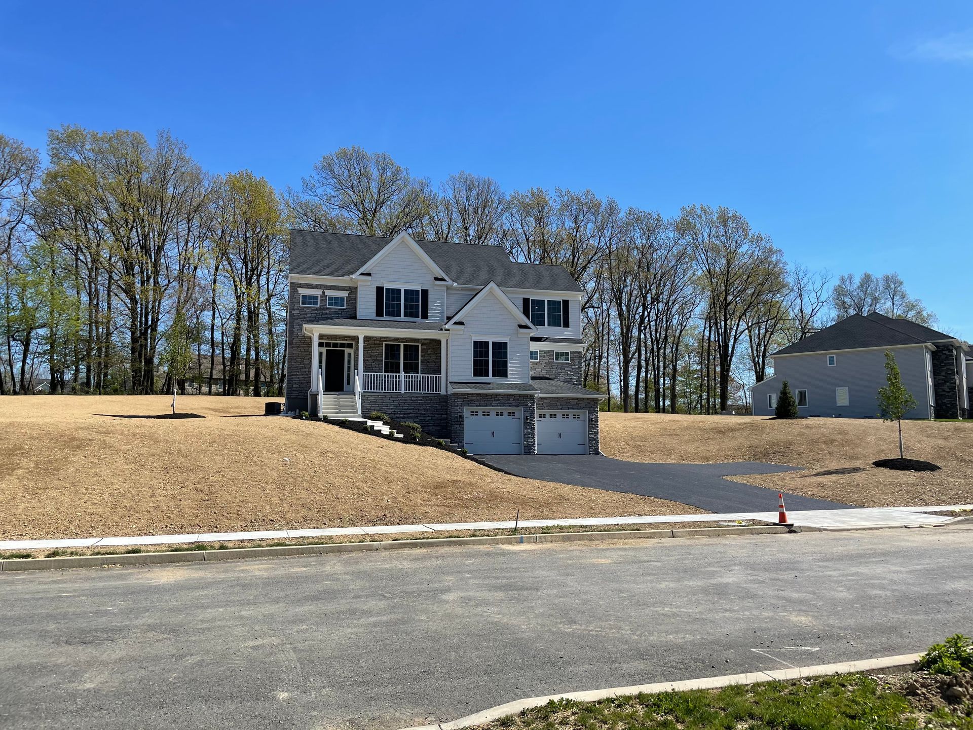 Two-story white house with gray roof and garage. Asphalt driveway slopes down to road. Construction site.