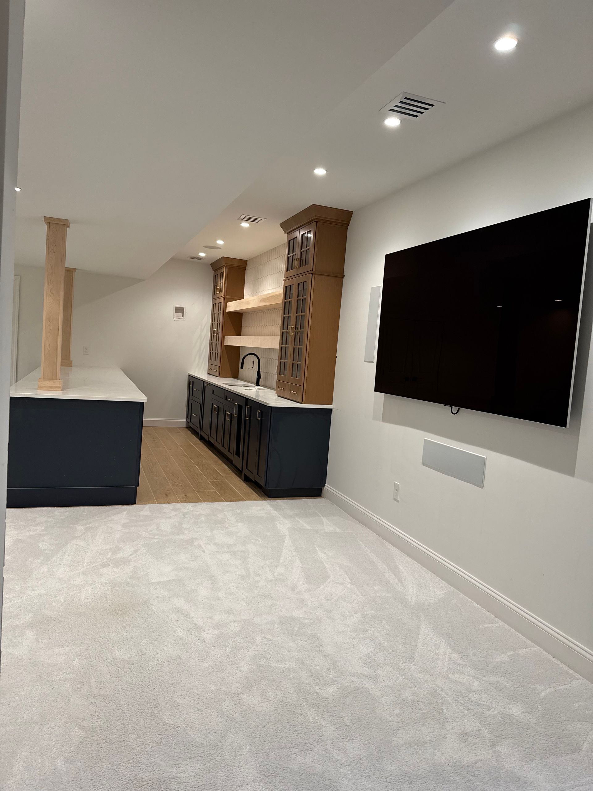 A modern home bar with dark blue cabinetry, light wood shelves, and a large TV on a white wall.