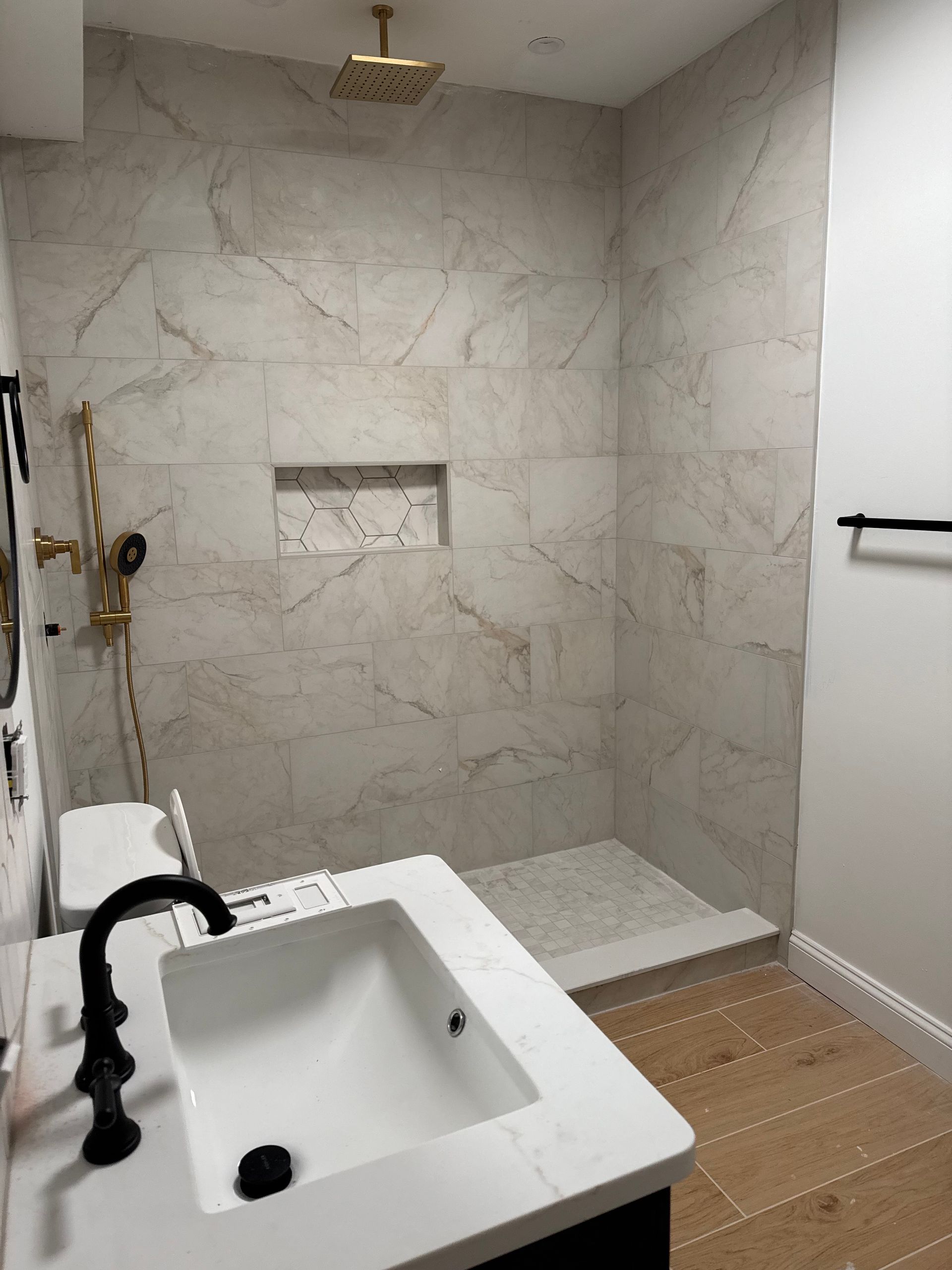 Modern bathroom with marble-tiled shower, gold fixtures, and black vanity. Wooden floor.