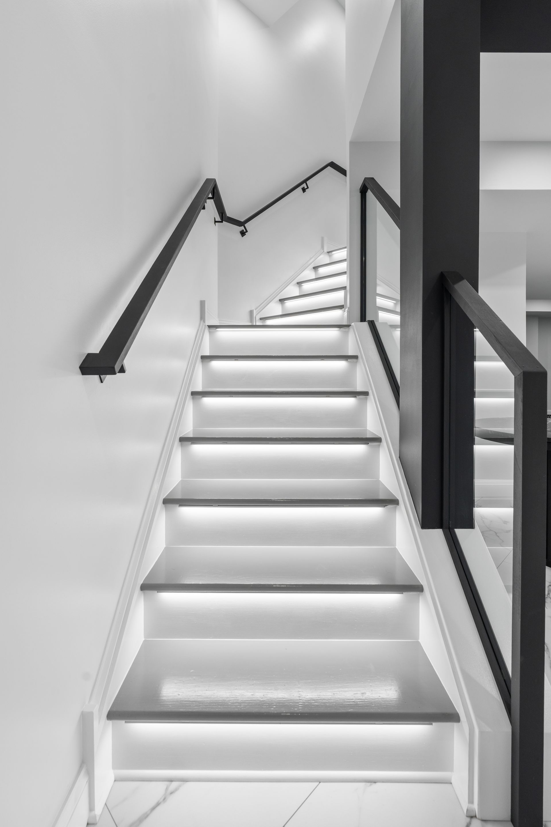Modern staircase with white steps, integrated LED lighting, and black handrails against white walls.