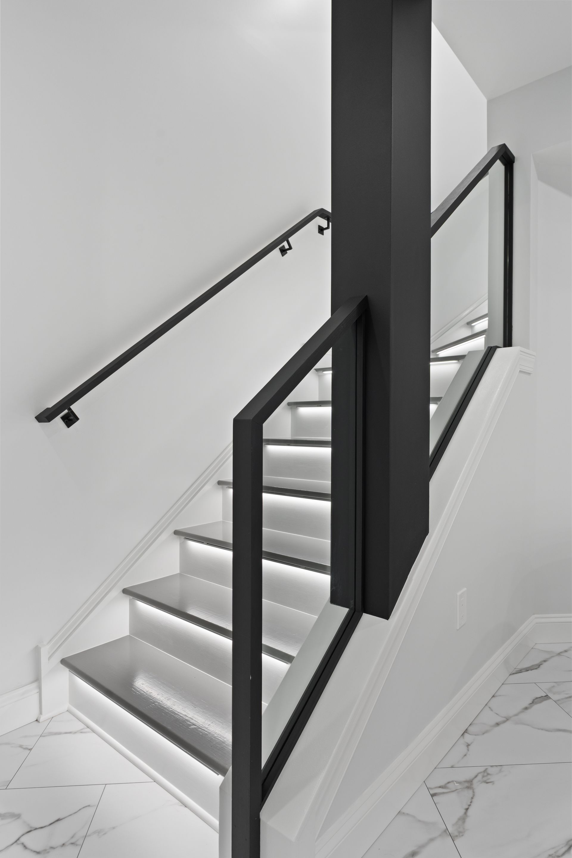 Modern indoor staircase with white marble floors, illuminated steps, a black support pillar, and matching black railings.
