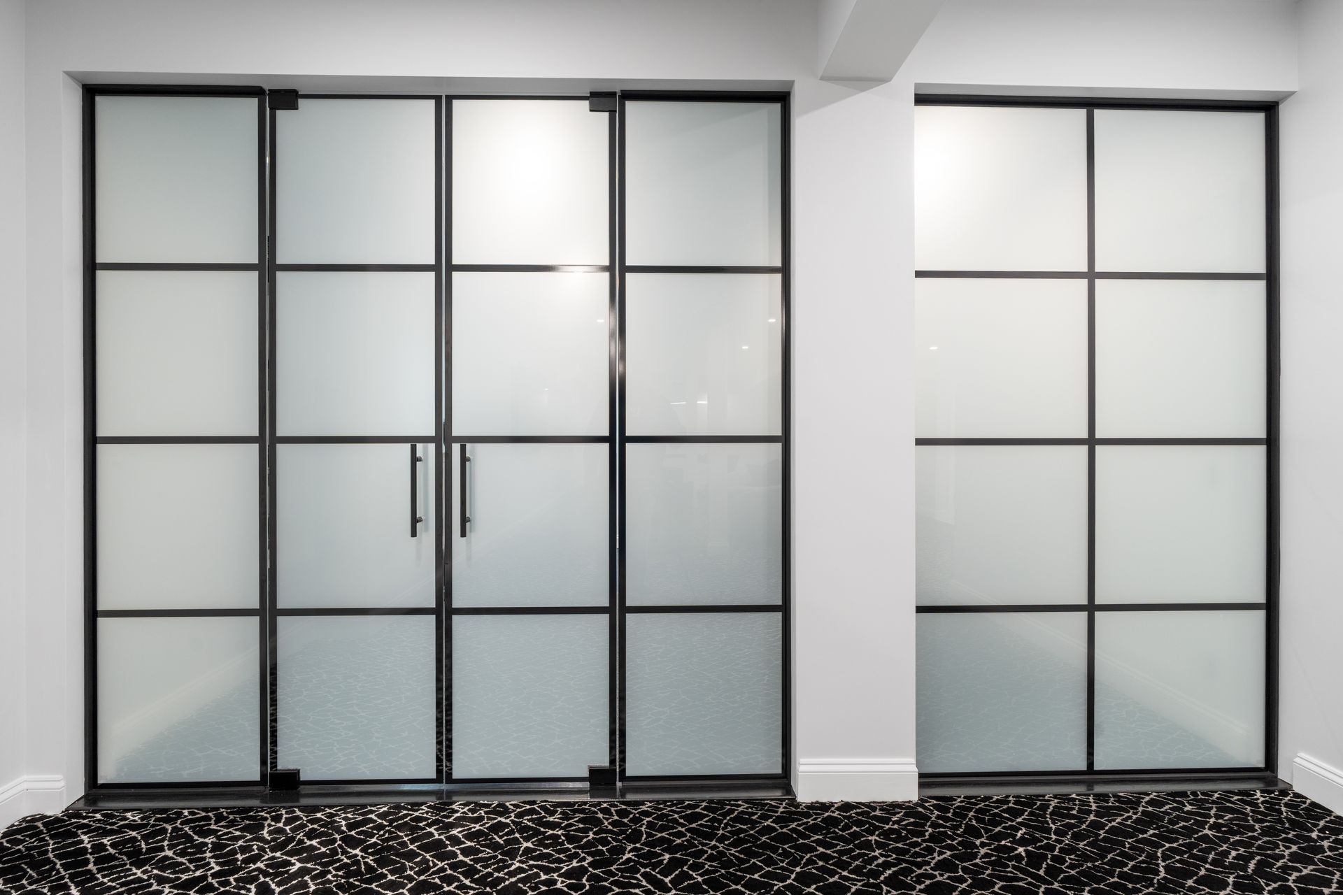 Modern black-framed glass partitions with frosted panels set against white walls above a patterned, dark floor.