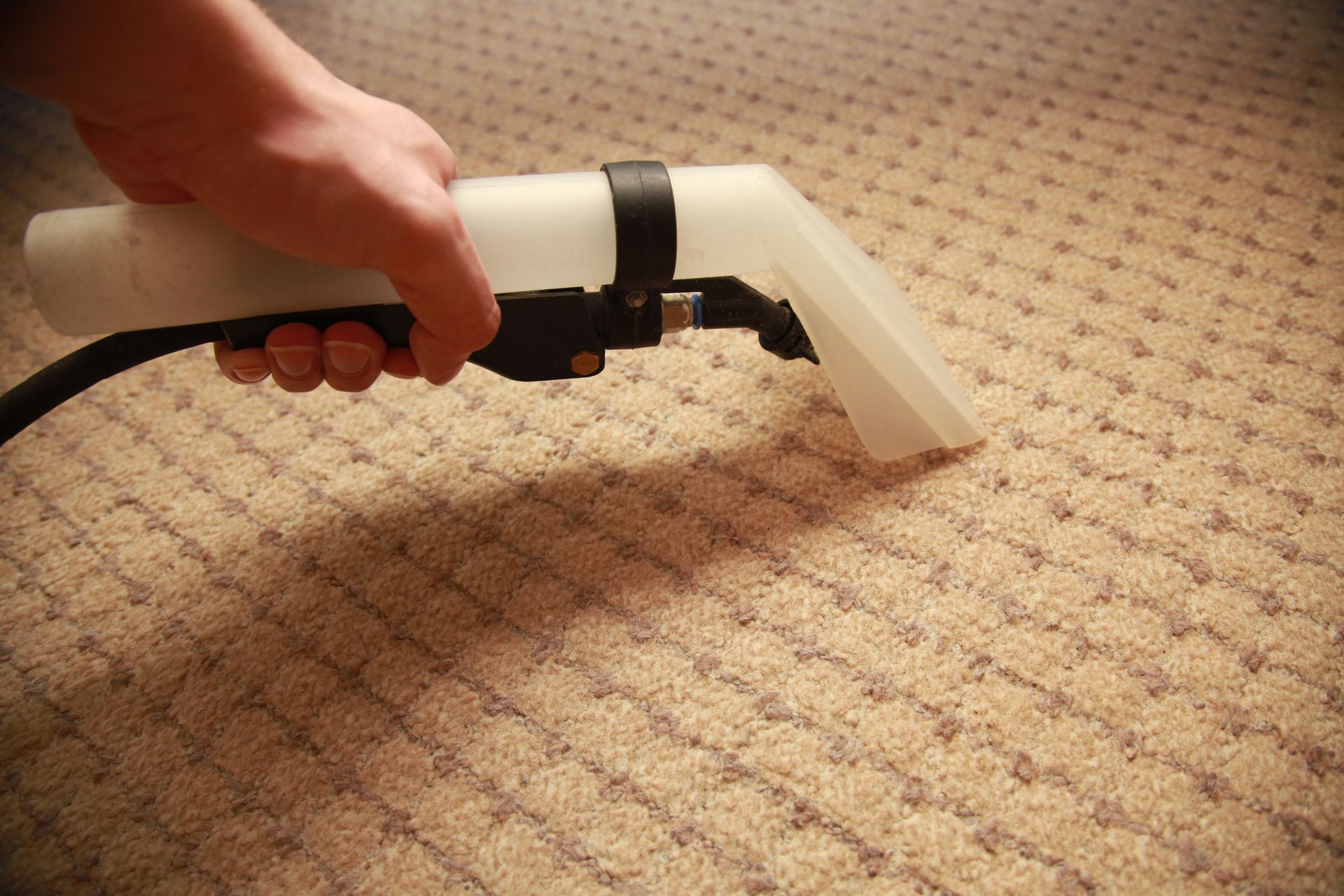 A hand uses a white upholstery cleaning tool to clean a beige, patterned carpet.
