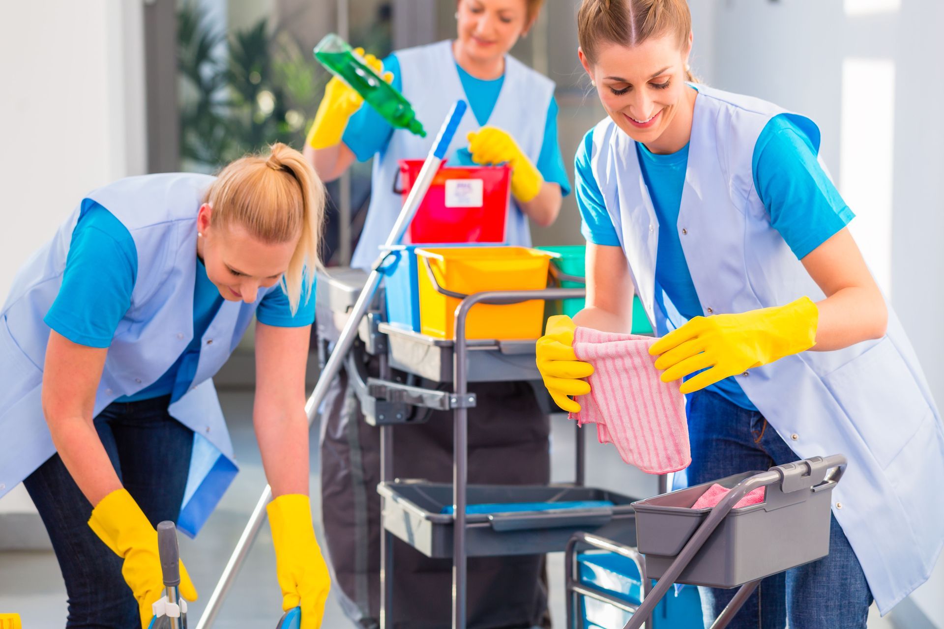 Three cleaners in blue uniforms and yellow gloves work in an office, using cleaning supplies and a utility cart.