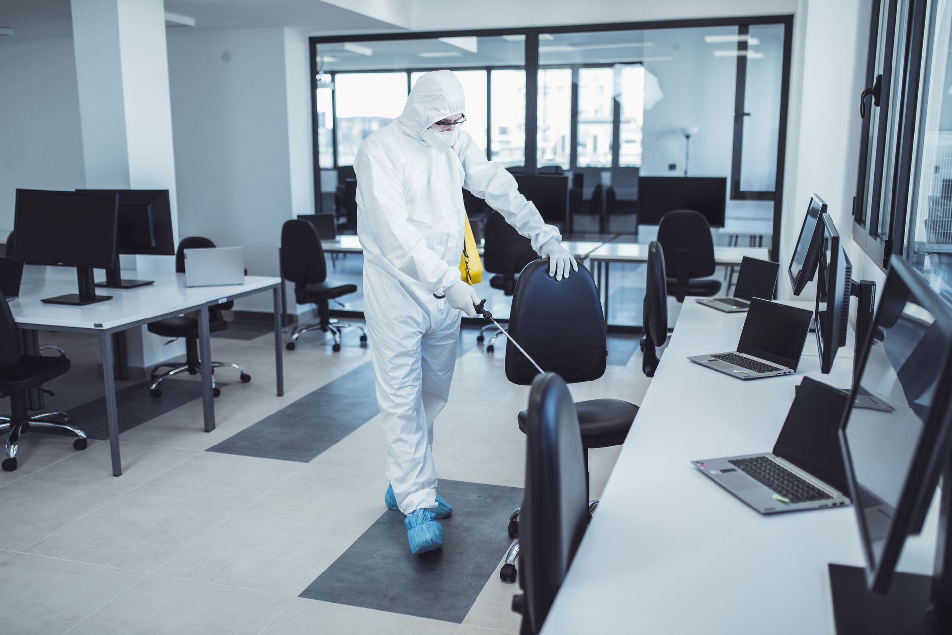 A person in a white protective suit and face mask sanitizes office chairs with a spray bottle in a modern office space.