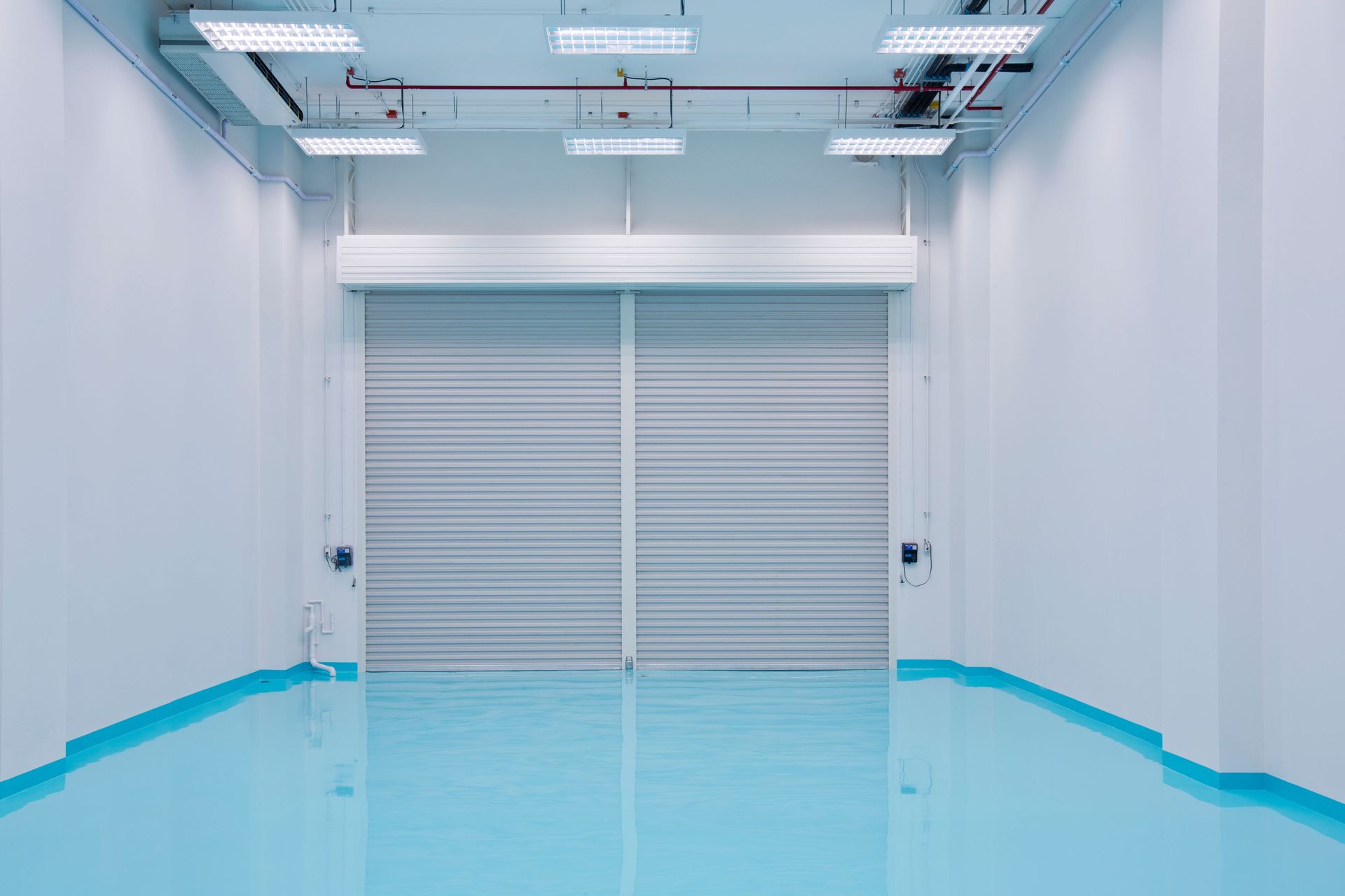 An empty, bright industrial room with white walls, a shiny blue floor, and a closed grey metal roll-up security door.