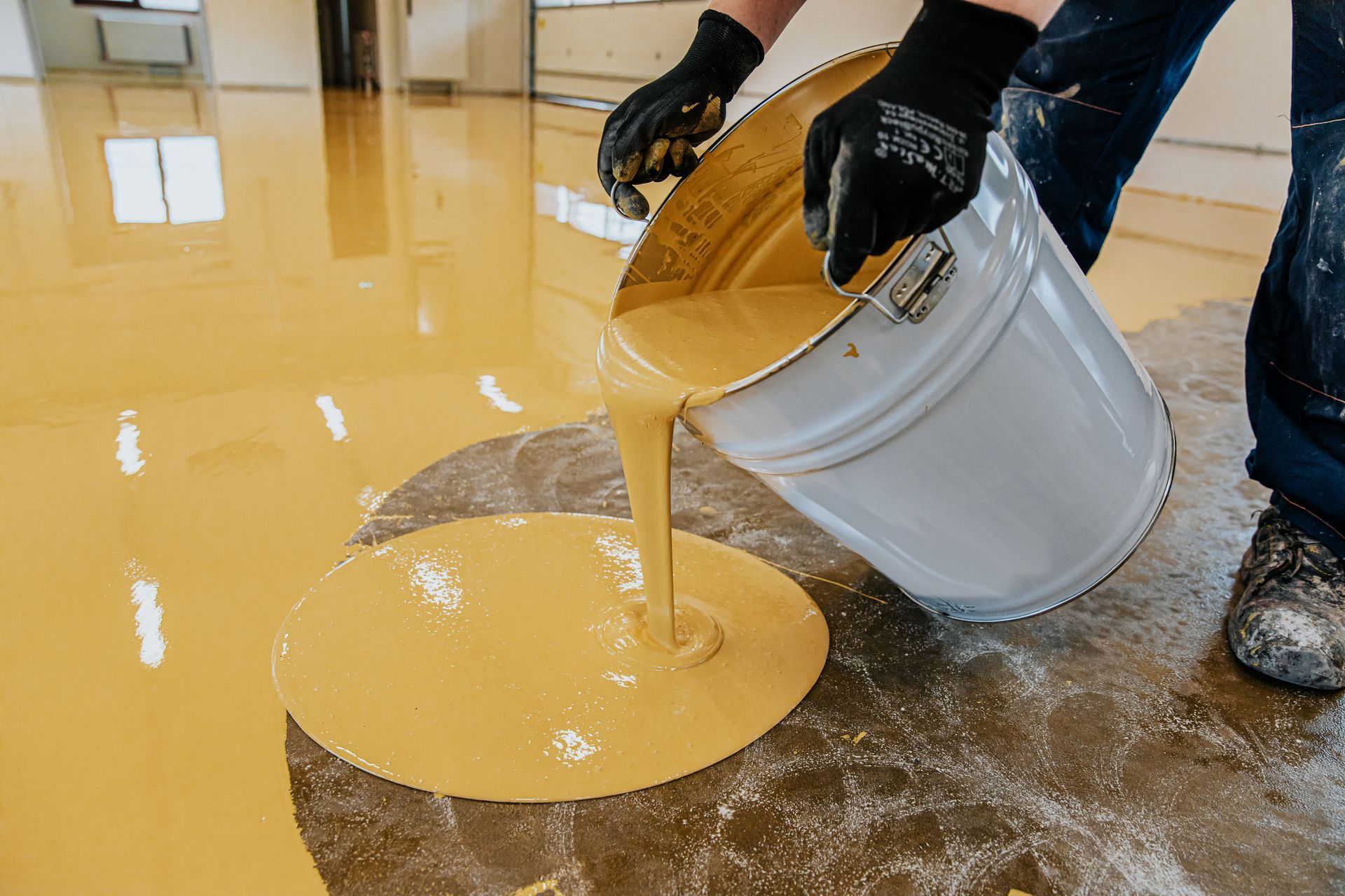 A person wearing work gloves pours thick, yellow epoxy resin onto a concrete floor.
