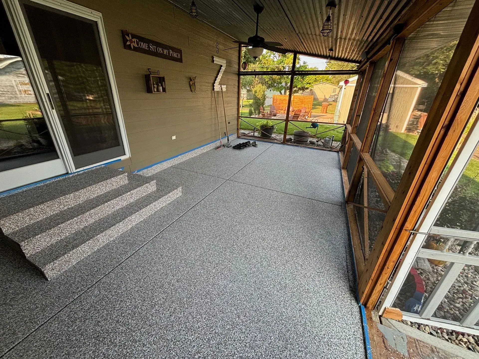 A covered patio with gray speckled flooring, two steps up to a sliding glass door, and a screen wall looking onto a yard.