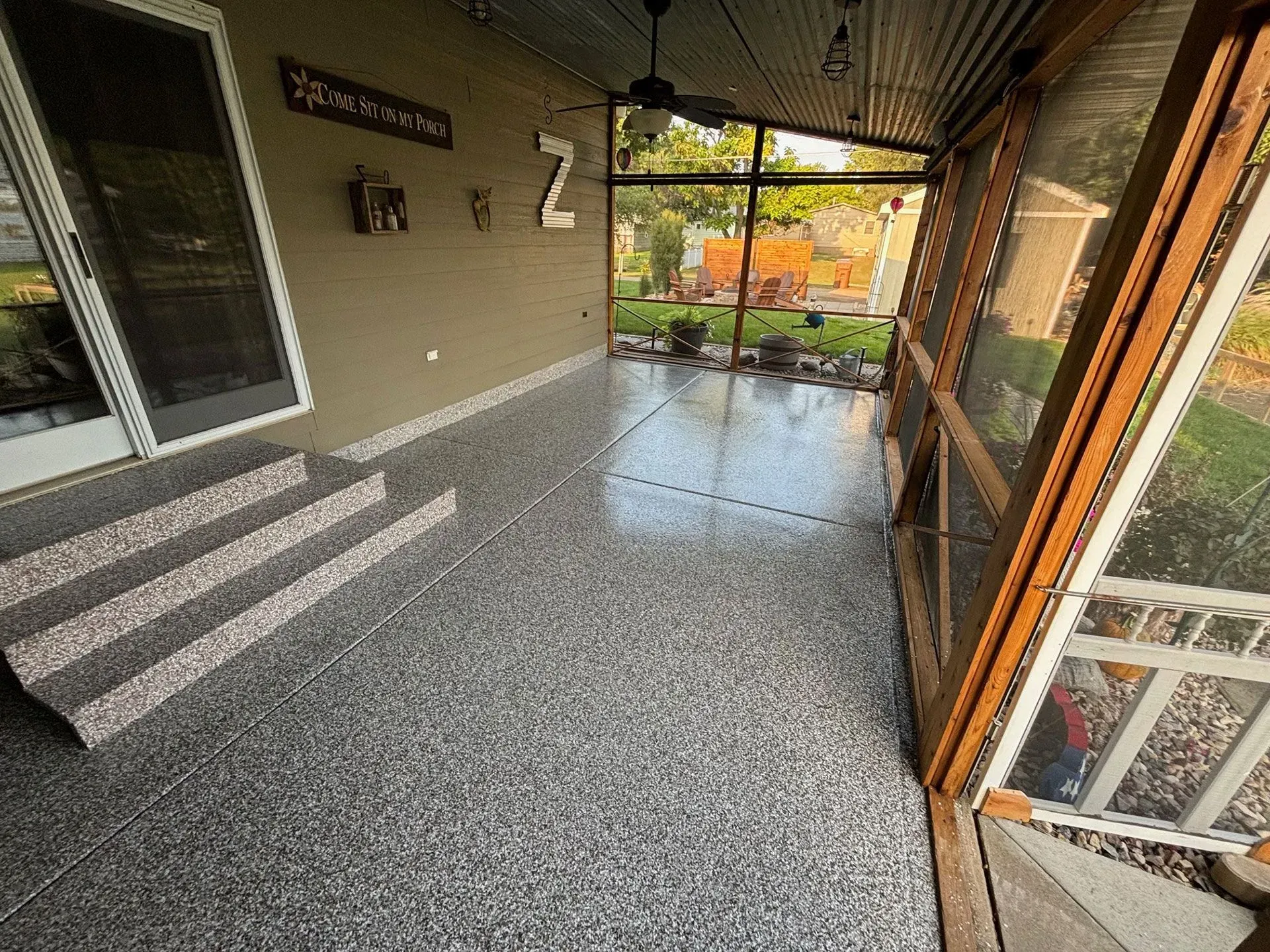 A screened-in porch with gray speckled flooring, stairs leading to a sliding door, and a ceiling fan.