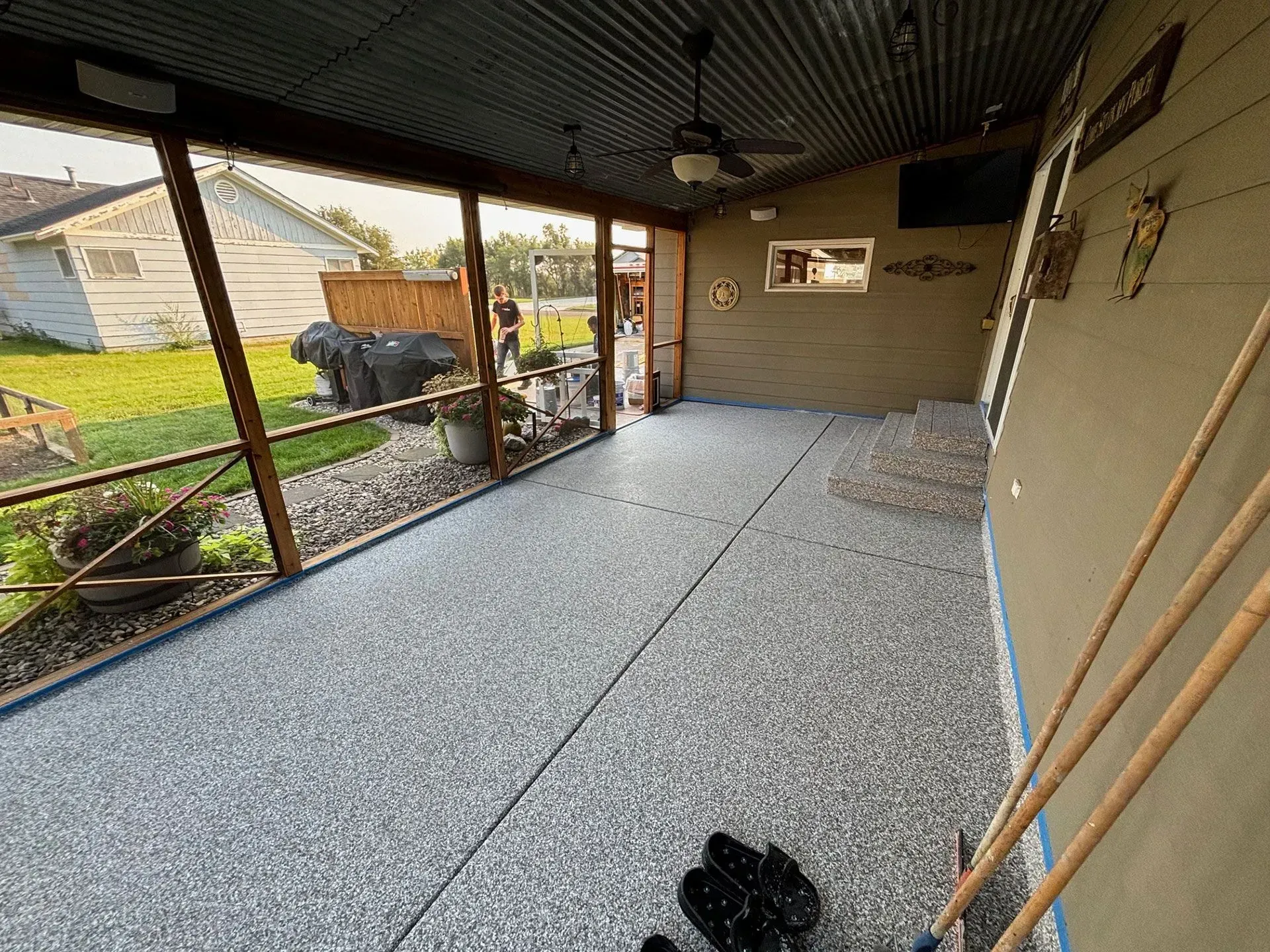 A covered outdoor porch with a speckled grey concrete floor, a ceiling fan, and screened walls overlooking a yard.