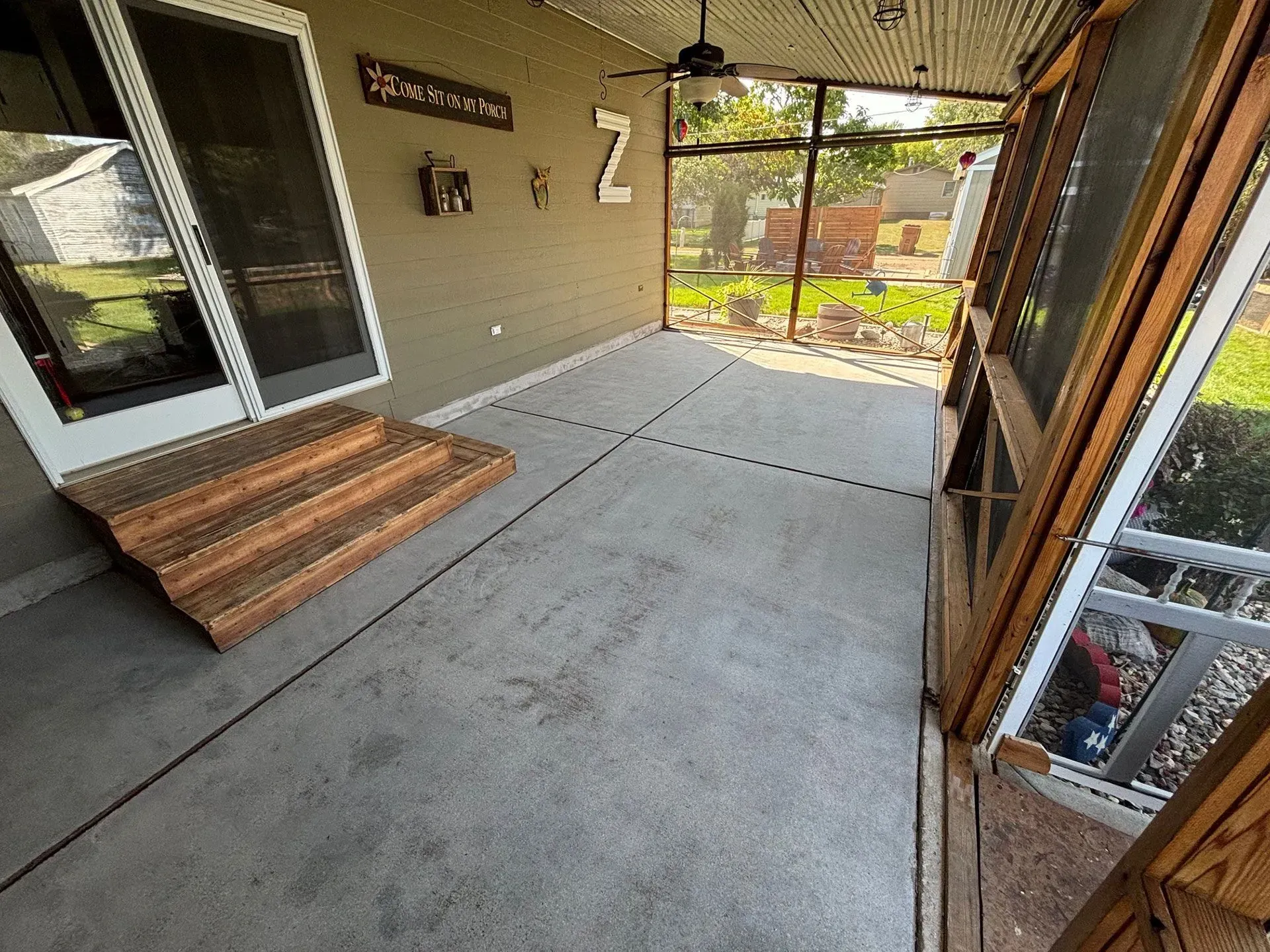 A covered patio with a concrete floor, a wooden step leading to a sliding glass door, and screened walls.