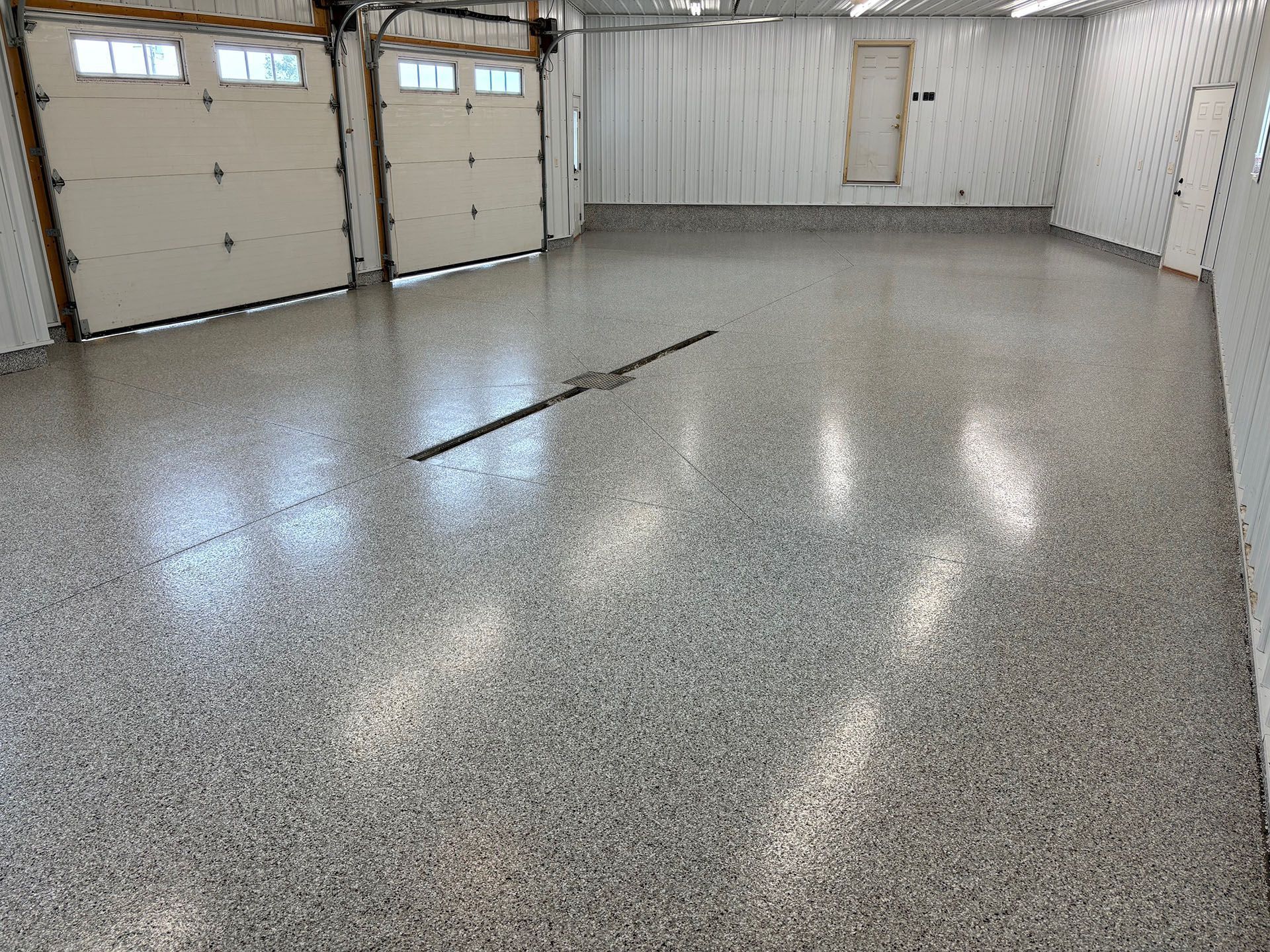A wide view of a spacious garage featuring a glossy, grey, speckled epoxy floor and two white garage doors.
