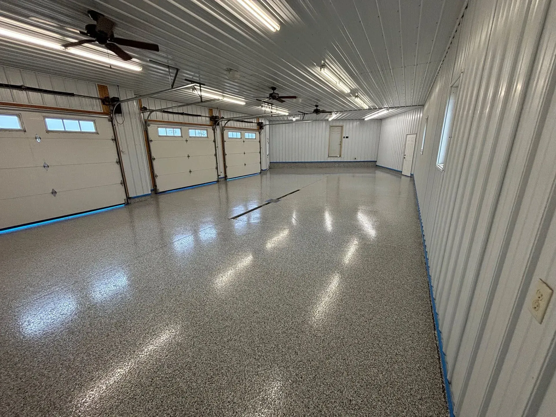 A spacious, empty garage with white metal walls, multiple white doors, ceiling fans, and a speckled epoxy floor.