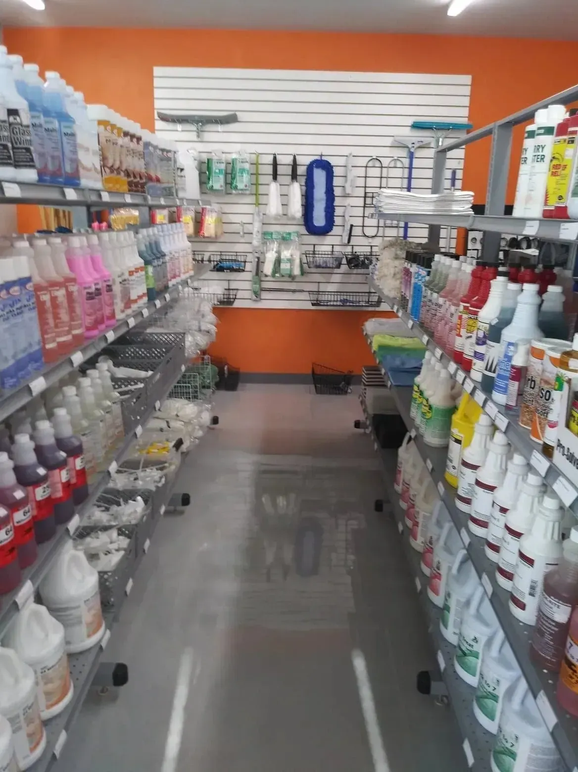 An aisle in a store with shelves packed with cleaning supplies against a bright orange wall.