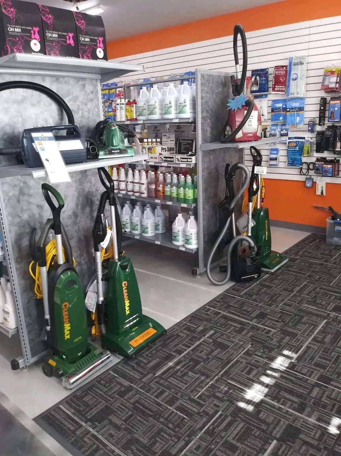 A retail store aisle featuring rows of upright and canister vacuums on shelves and the floor, with cleaning supplies.