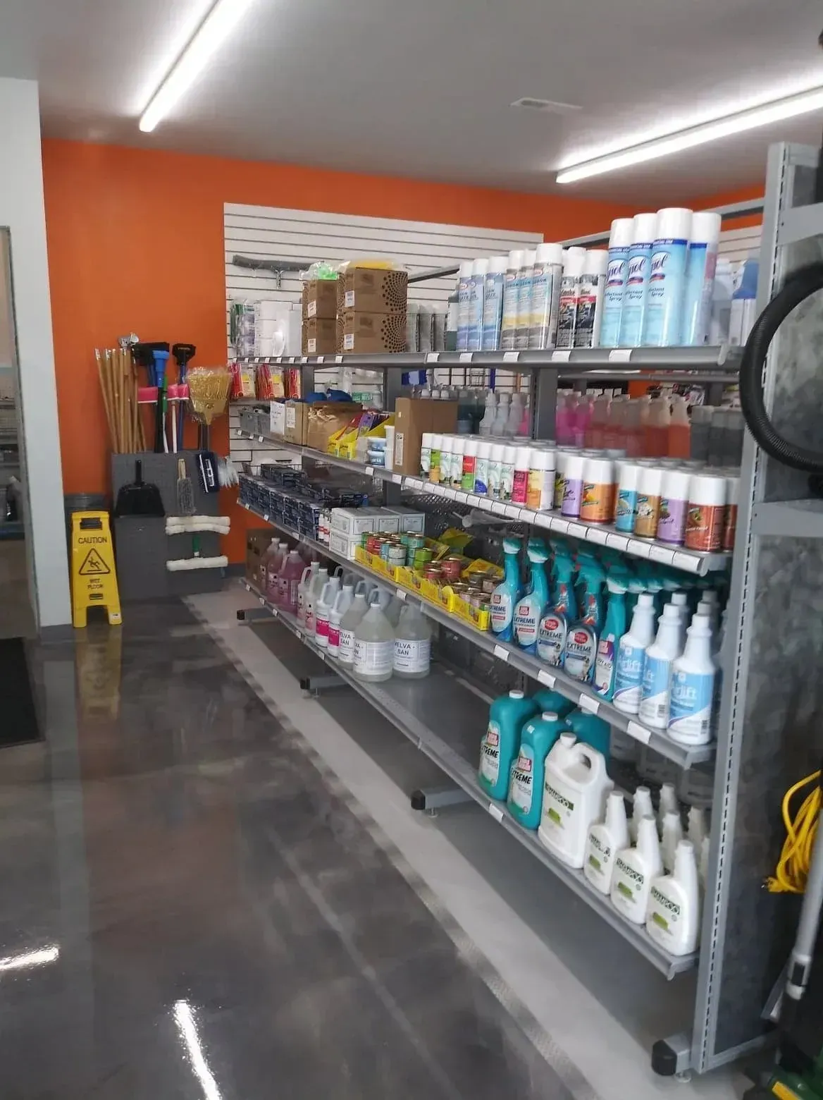 Retail shelving stocked with cleaning supplies and household goods, set against a bright orange wall.