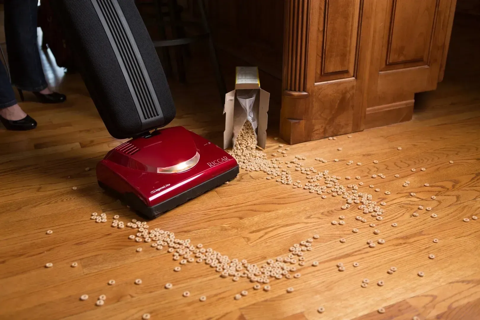 A red vacuum cleaner approaches a pile of cereal spilled from a box onto a hardwood floor.