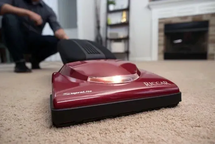 A close-up of a maroon Riccar vacuum cleaner on a carpeted floor, with a person crouching in the blurred background.