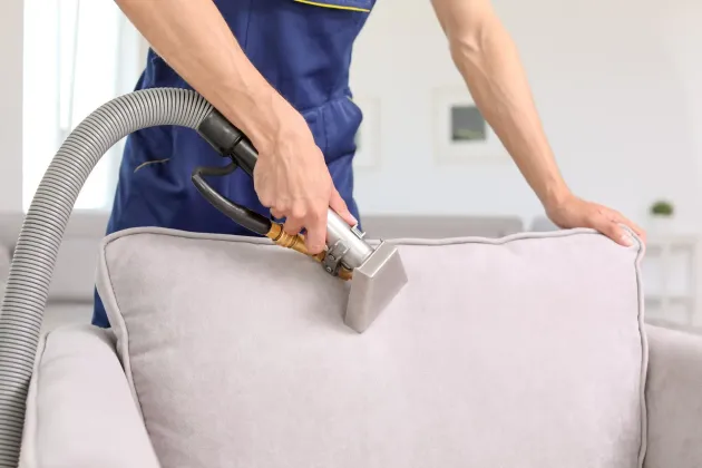 A person in a blue uniform uses a professional steam cleaner to clean the fabric of a light gray armchair.