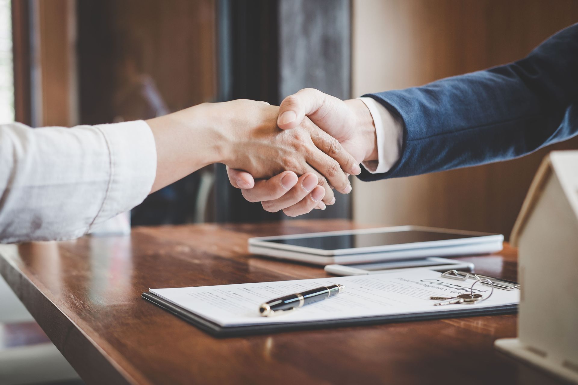 Two people shake hands over a desk with a contract and a model house, signaling a real estate or business agreement.