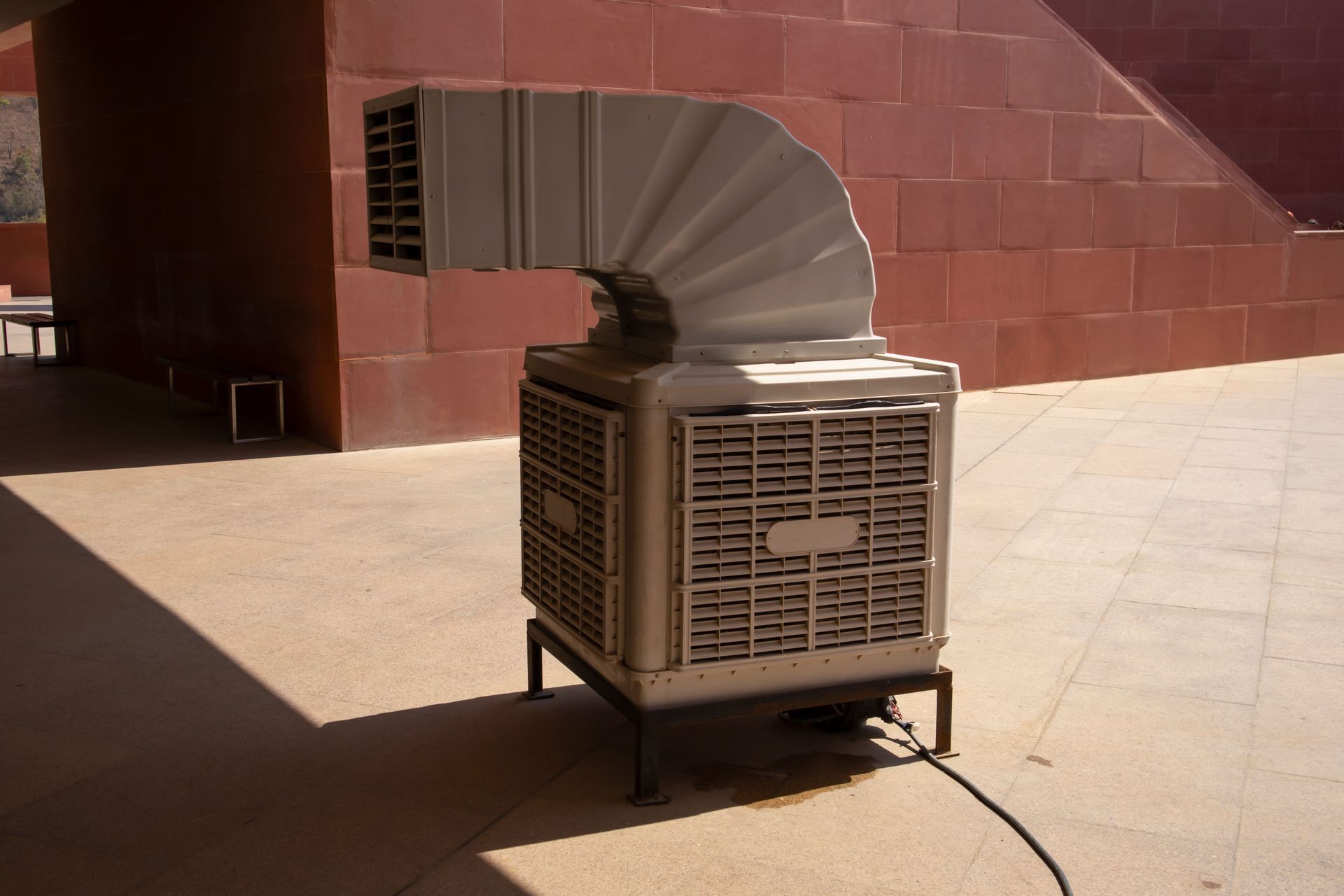 A beige evaporative air cooler with an attached flexible duct stands on a paved patio against a red brick wall.