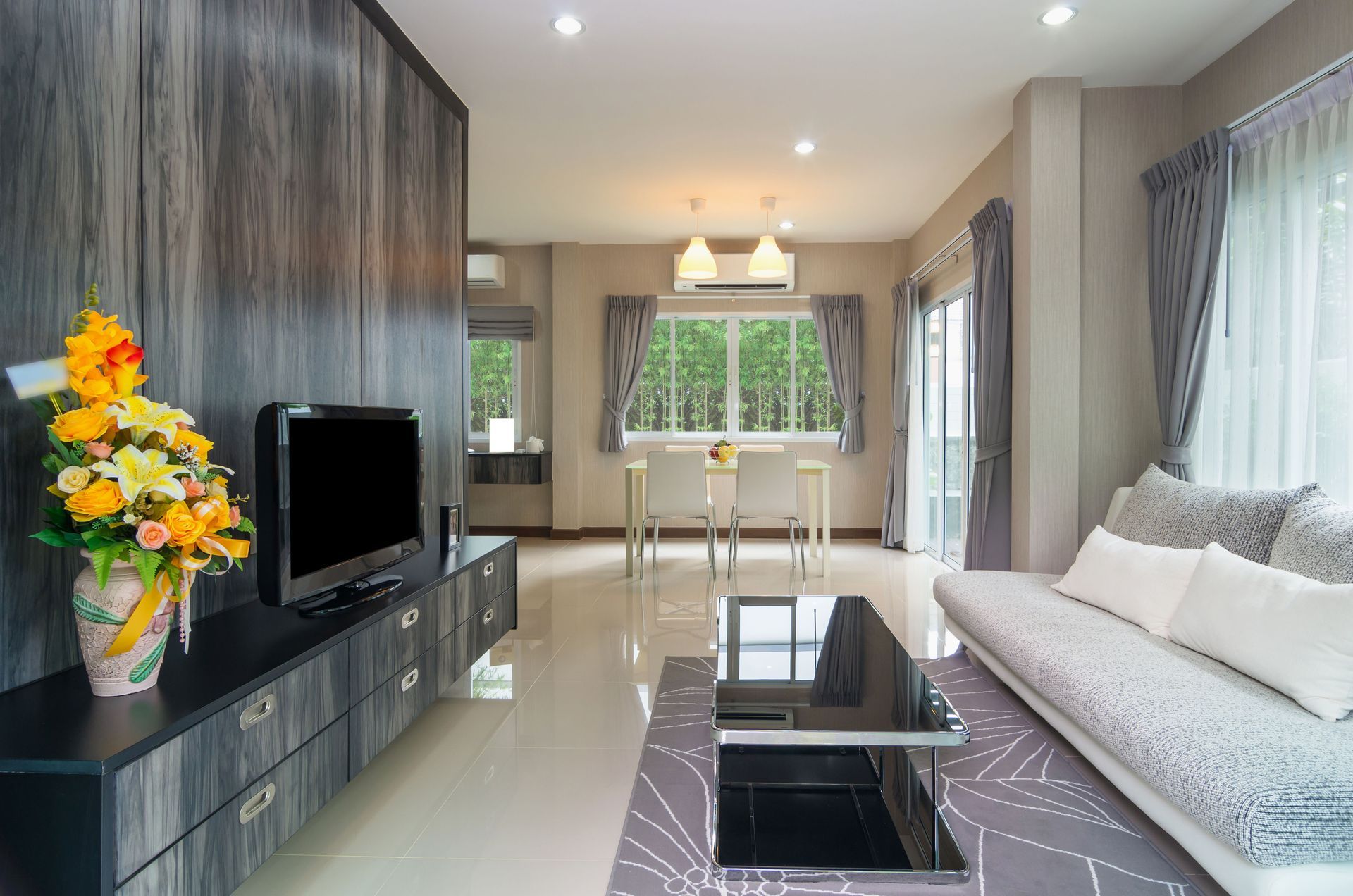 Modern living room with a gray wood-paneled TV wall, sofa, glass coffee table, and a dining set near a large window.