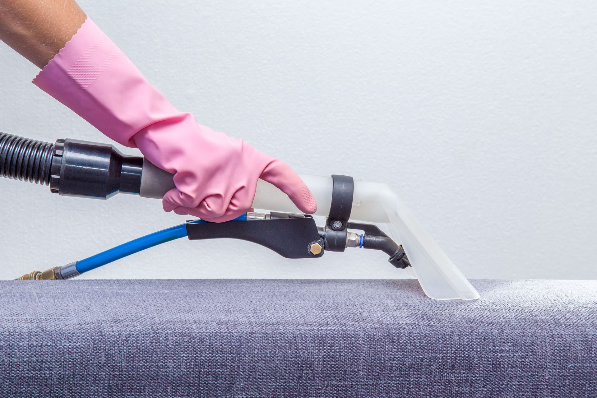 A hand in a pink glove uses a carpet cleaning vacuum attachment to clean a grey fabric surface.