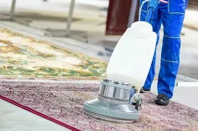 A person in blue workwear uses a carpet cleaning machine on a patterned rug in a facility.