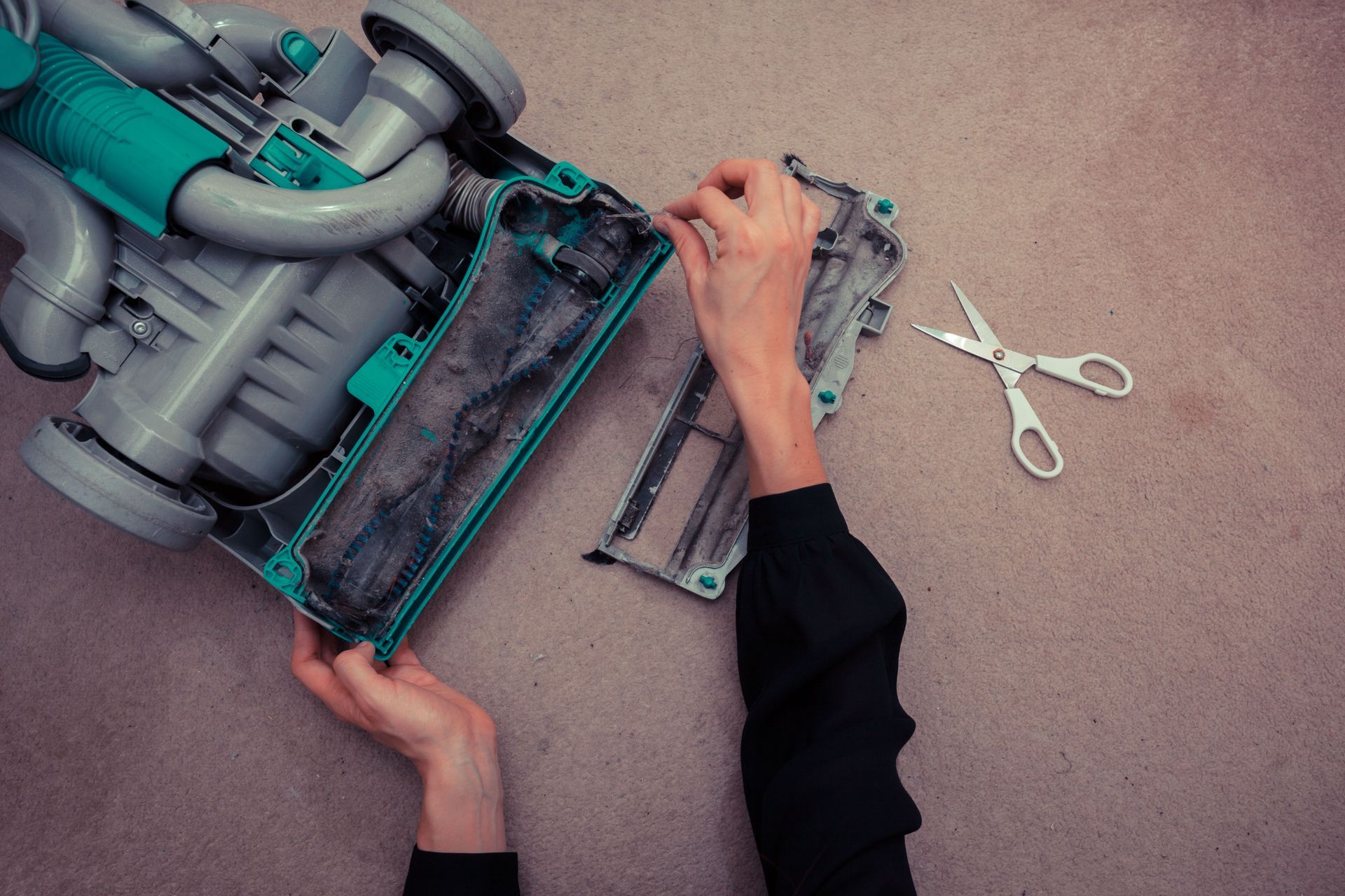 Hands cleaning hair and debris from a vacuum cleaner brush roll with scissors nearby on a carpeted floor.
