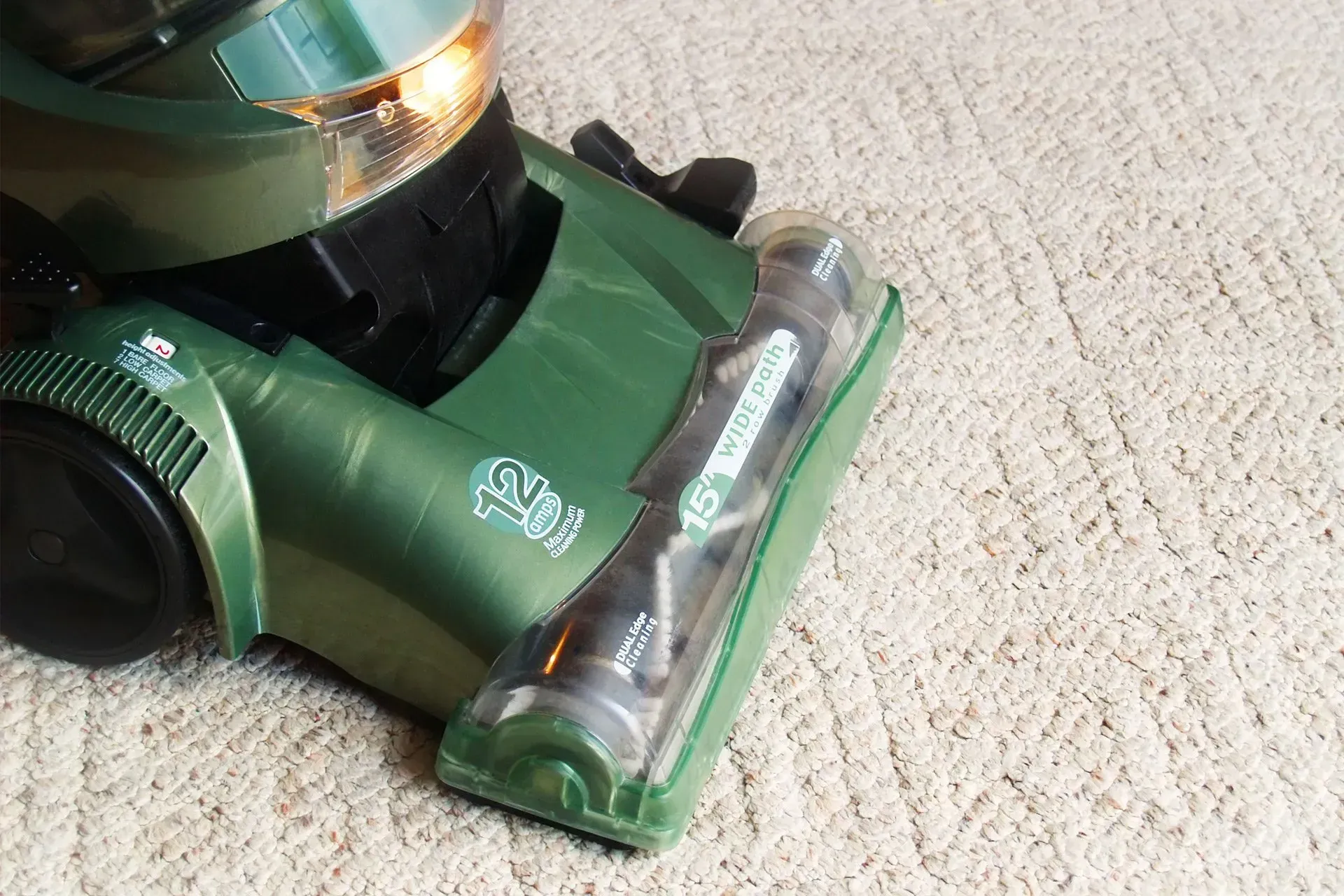 A green vacuum cleaner sits on beige carpet, showing a clear brush roll housing and a headlight turned on.