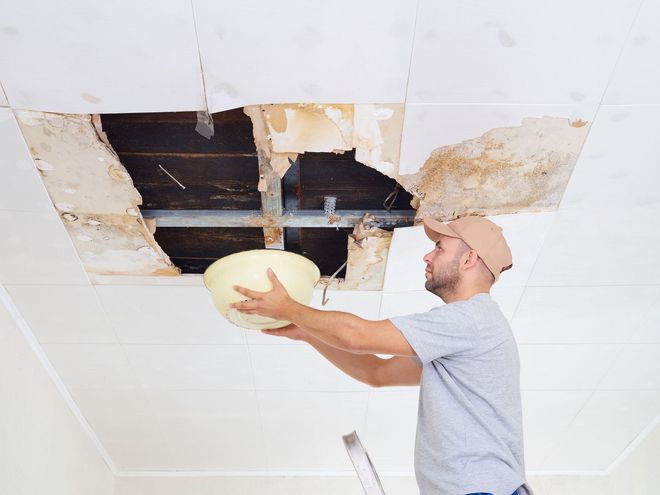 A person on a ladder holds a bowl to catch water leaking through a large, damaged hole in a white ceiling.