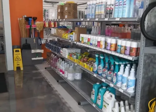 Shelves in a store aisle stocked with various cleaning supplies, bottles, and a yellow caution floor sign.